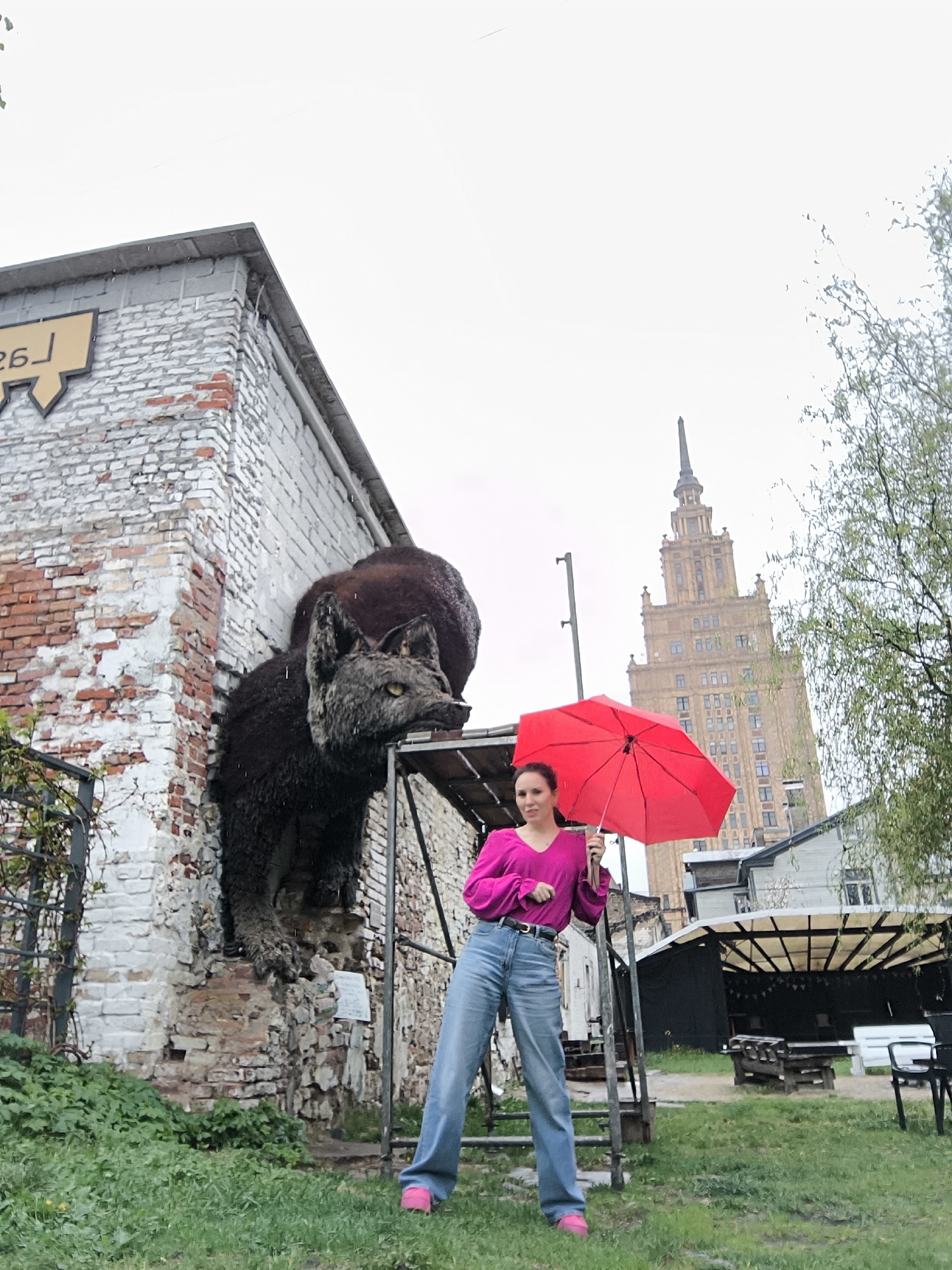 A medium shot captures a woman standing on a gray metal structure in front of a brick building with an animal sculpture attached to the wall. The woman has fair skin and dark hair tied back. She wears a bright pink blouse, high-waisted blue jeans, and pink shoes, and holds a closed red umbrella.
The building is composed of reddish-brown and white bricks. The animal sculpture, featuring a dark wolf-like head and a large brown body, appears to be emerging from the wall. To the left of the building, vegetation grows on the ground. Behind the woman and to the right, there is a distinctive building with a spire, identifiable as the Latvian Academy of Sciences in Riga, Latvia. Further to the right is a partially obscured outdoor structure with a black exterior and a white roof. The background is overcast.