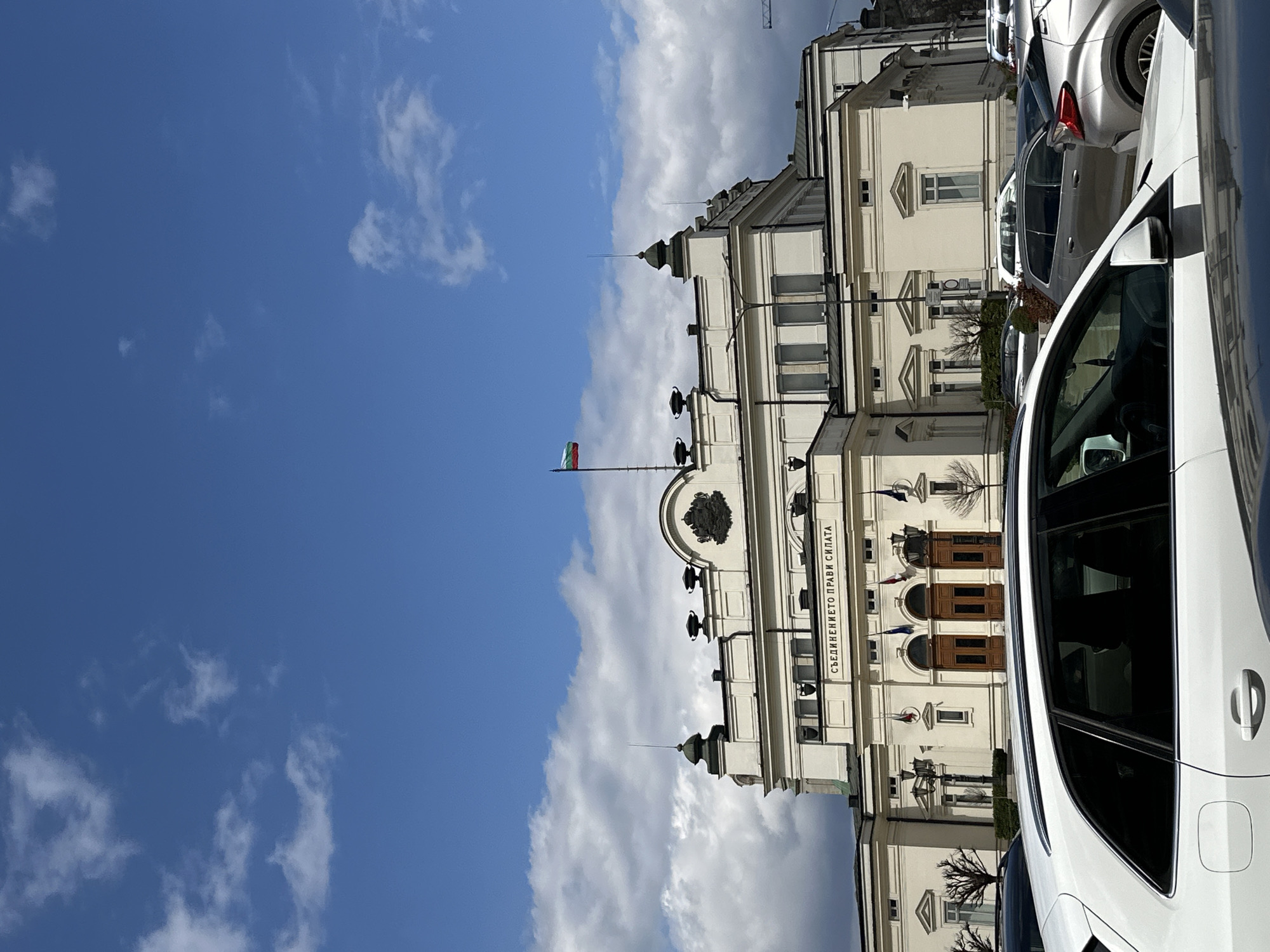The image shows the National Assembly of Bulgaria in Sofia on a sunny day with blue sky and clouds. The building is cream-colored with a Bulgarian flag on top. Parked cars are visible in front. The inscription “Съединението прави силата” (Unity makes strength) is displayed on the building.