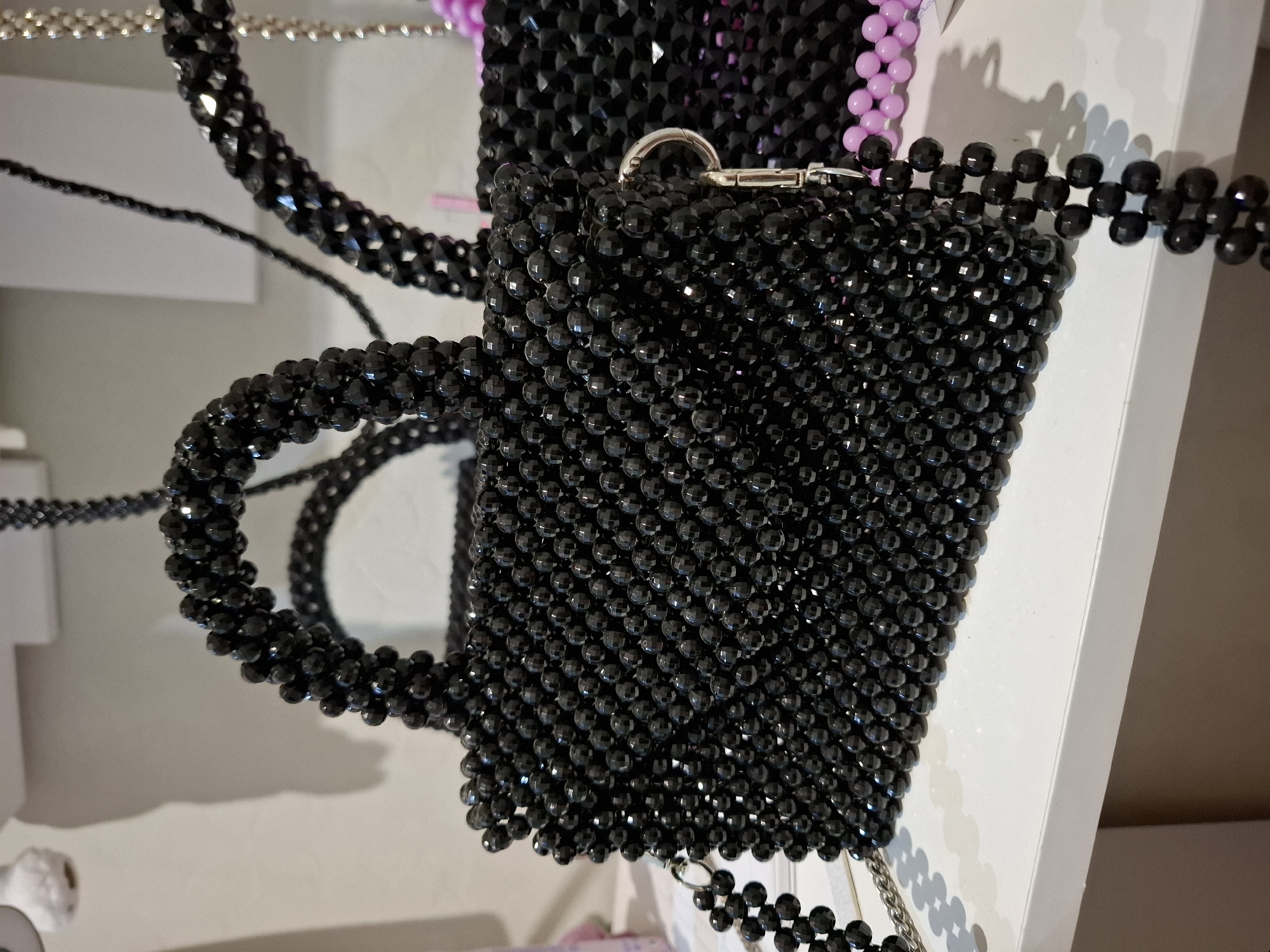 The image shows a close-up of a black beaded mug-shaped handbag. The mug is entirely covered in small, faceted black beads, giving it a textured and shiny appearance. It has a handle also made of black beads, curved to mimic a mug's handle. A silver clasp is visible at the top of the mug, suggesting it might have a lid or closure.
In the background, parts of other beaded items are visible, including a chain made of black beads and a section of a bag or accessory made of light pink beads. The items appear to be displayed on a white surface, possibly a shelf or table. The lighting suggests an indoor setting, with soft shadows cast on the surface. No people are visible in the frame. The image is focused on the handbag, highlighting its craftsmanship and material. There is no visible text in the image. The overall impression is of a decorative and perhaps handmade accessory.
