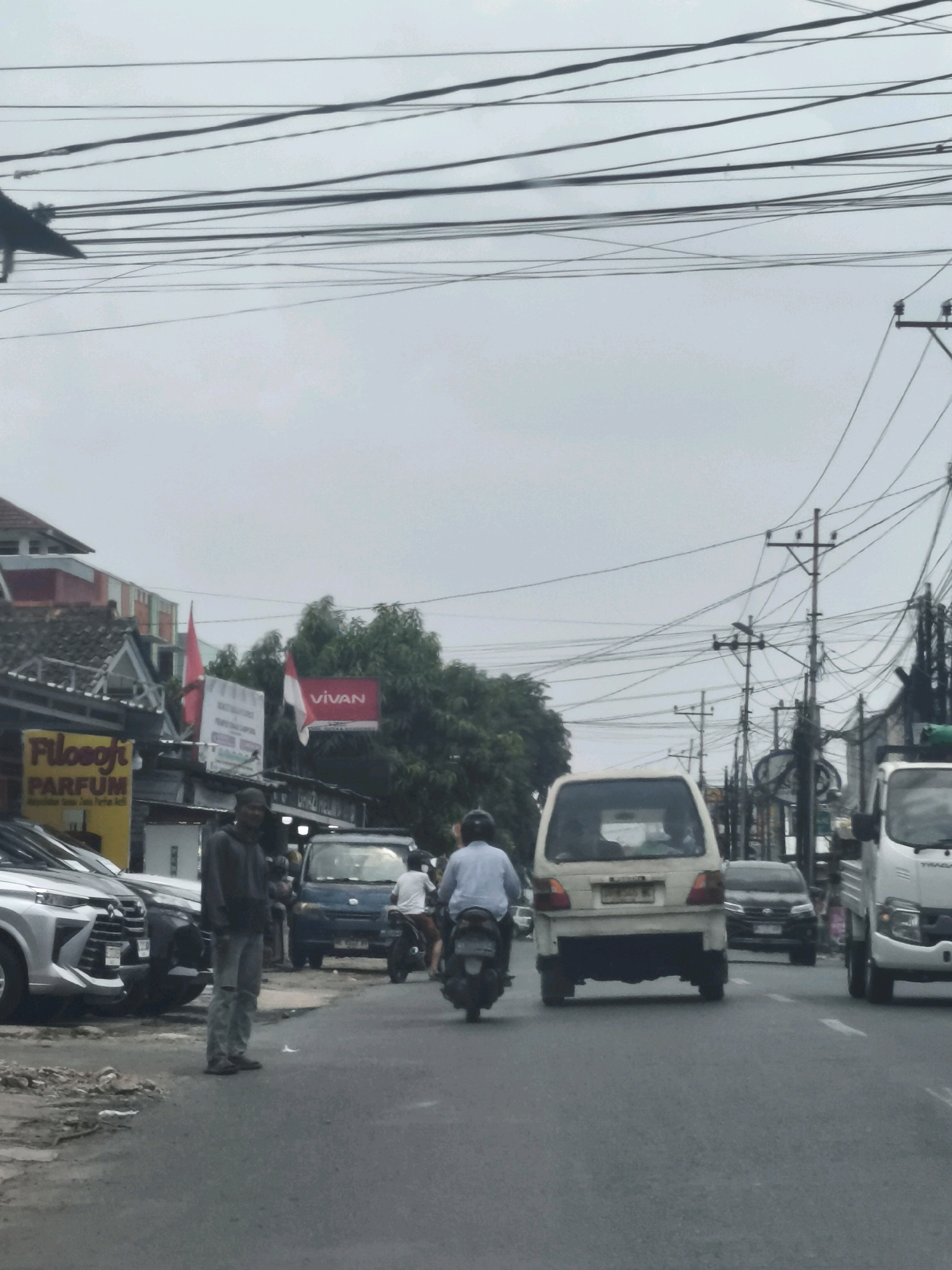The image depicts a street scene in Bandar Lampung, Indonesia, characterized by heavy overhead utility wires. On the left side of the road, a storefront displays a yellow sign with "Filosofi PARFUM" written in black. Further down the street, a man in a dark hoodie and ripped jeans stands on the roadside. A silver SUV is parked to his left, and a blue van is visible behind him. A person on a motorcycle is proceeding along the road. In the center of the frame, a white van is driving, and further ahead, a white truck is also on the road. The sky is overcast.
