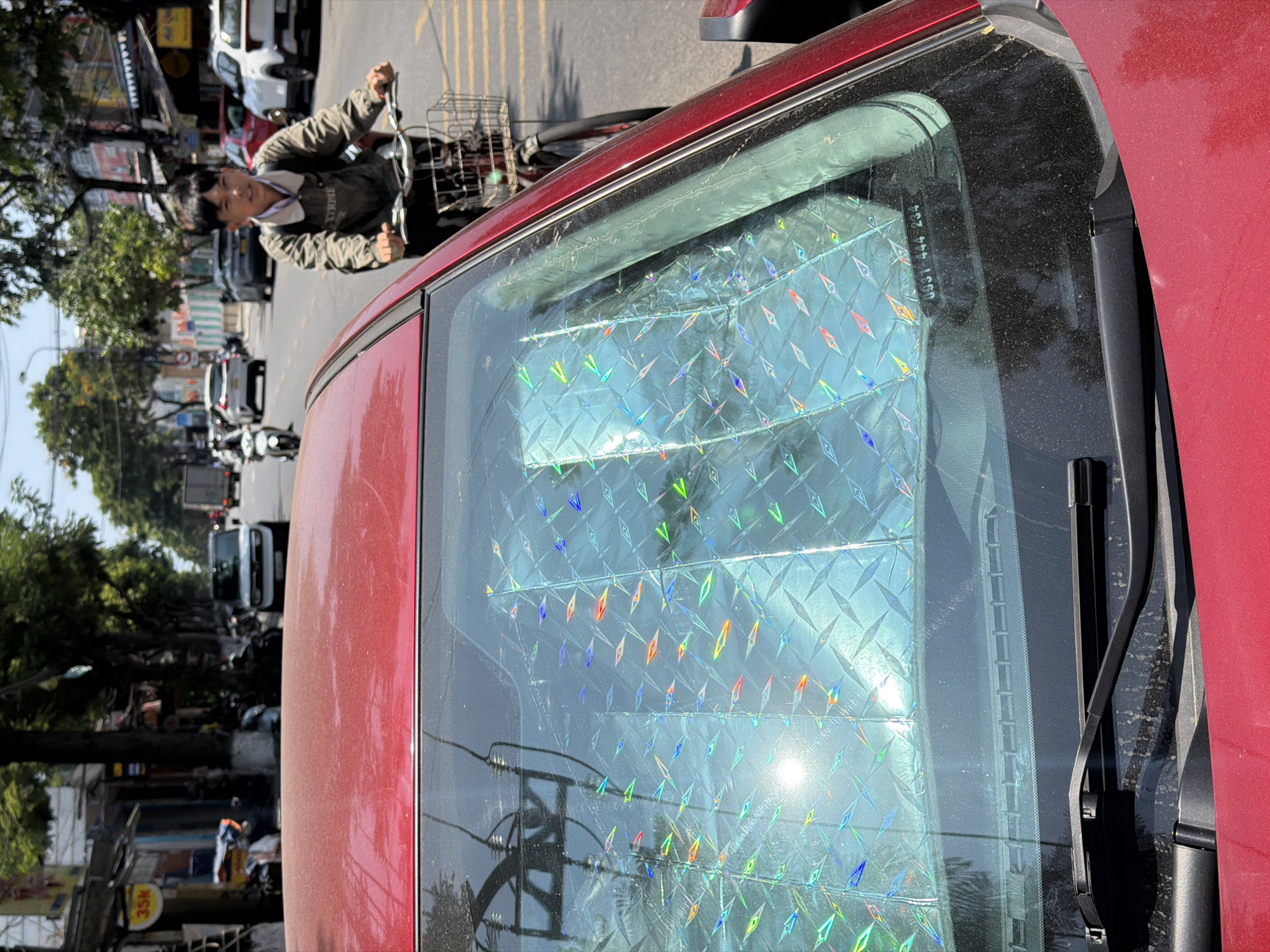 The image shows a red car with a holographic sunshade covering the rear window. A man on a bicycle is visible through the window, riding on a street lined with trees and buildings. Other vehicles are present on the road. The location is likely Đà Nẵng, Vietnam, based on street features and vehicle types.