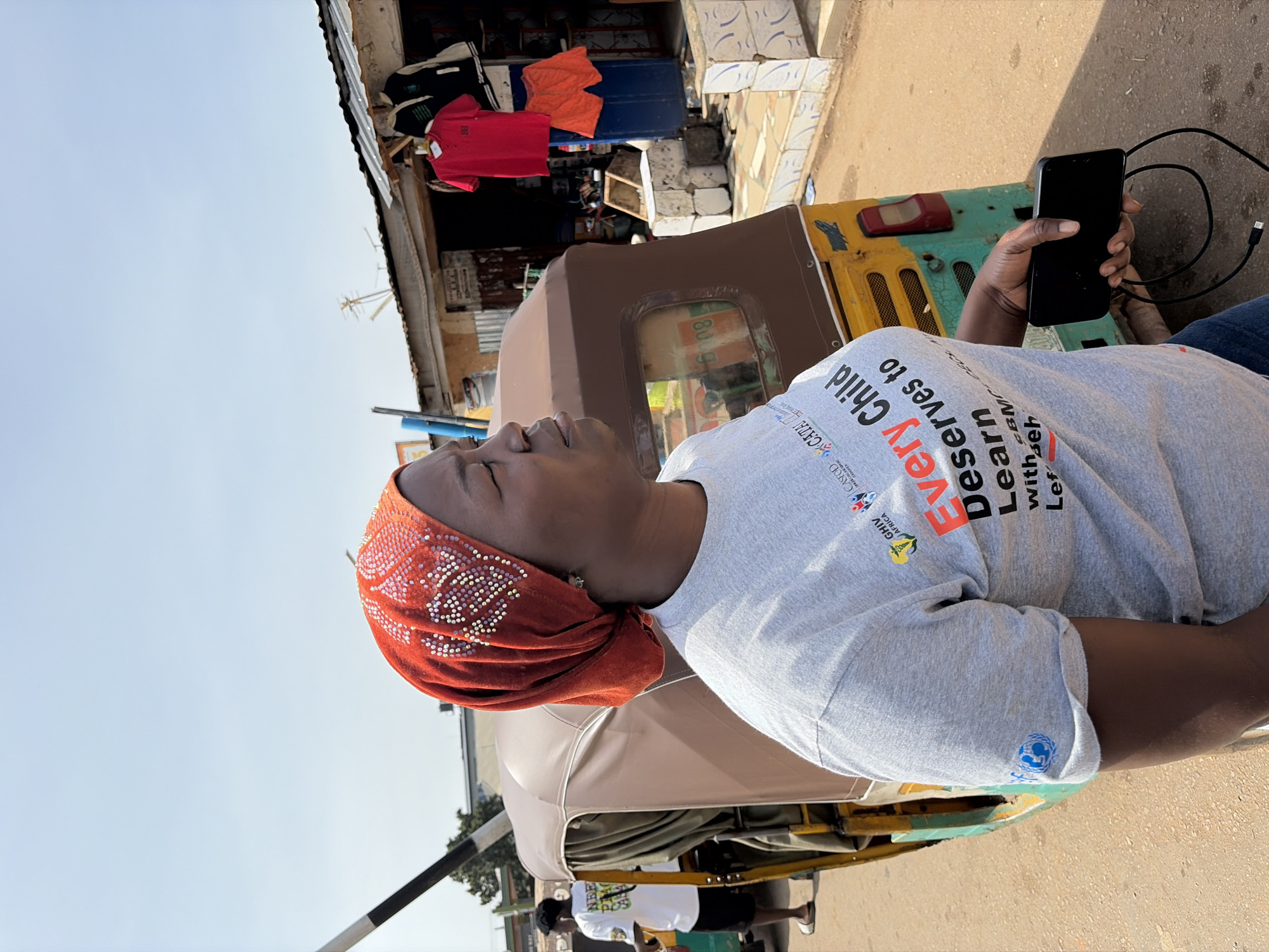 A bright, outdoor photograph captures a person in Gura Top, Nigeria, under a clear blue sky. The main subject is a person, likely a woman, with dark skin, wearing an orange headscarf decorated with white and pink beaded patterns. She is dressed in a light gray t-shirt and is looking upwards with a neutral expression. In her right hand, she holds a black smartphone connected to a black charging cable.
She appears to be seated next to or in a brown-canopied, three-wheeled vehicle, resembling a tuk-tuk, which has yellow and green sections on its rear. The background reveals a street or market scene with a building or stall where clothes, including red and orange shirts, are displayed. Another person in a white t-shirt is partially visible in the distance. The ground is a light-colored pavement, suggesting a dry, sunny day.
The gray t-shirt prominently features text: "Every Child Deserves to Learn with BMC Lef Beh", "GHIV AFRICA", and "CASFOD CATAL", along with several small logos, one of which appears to be a globe with human figures. Partially visible text "POS 608" is present on the vehicle. The overall scene depicts a vibrant, everyday setting in an urban or semi-urban environment.