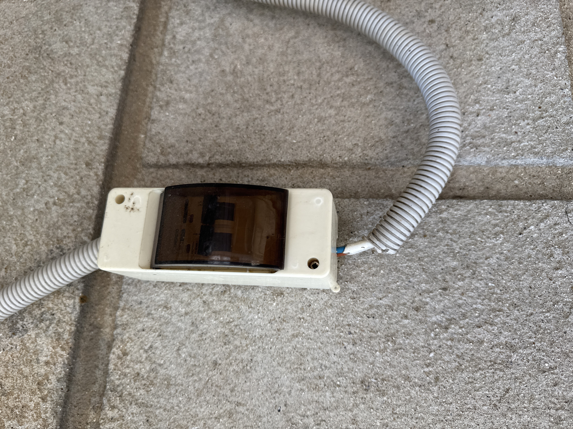 This media file features a close-up, eye-level shot of a cream-colored plastic electrical sensor with a dark, translucent cover. The sensor is mounted on a rough, gray concrete surface, possibly a sidewalk or patio, with visible grout lines. A ribbed, gray flexible conduit containing electrical wires emerges from the left side of the sensor, curving upwards and to the right. The wires, red and blue, are visible exiting the conduit and entering the sensor on the right side. There is a small, circular hole on the left side of the sensor, and another similar hole on the right side. The sensor appears to be a motion detector or a similar outdoor electrical component, given its placement on concrete and the protective conduit. The lighting suggests daytime, but the weather is not discernible. No people are visible. There is no visible text in the image. The context provided indicates the location is Rozdilna, Ukraine.
