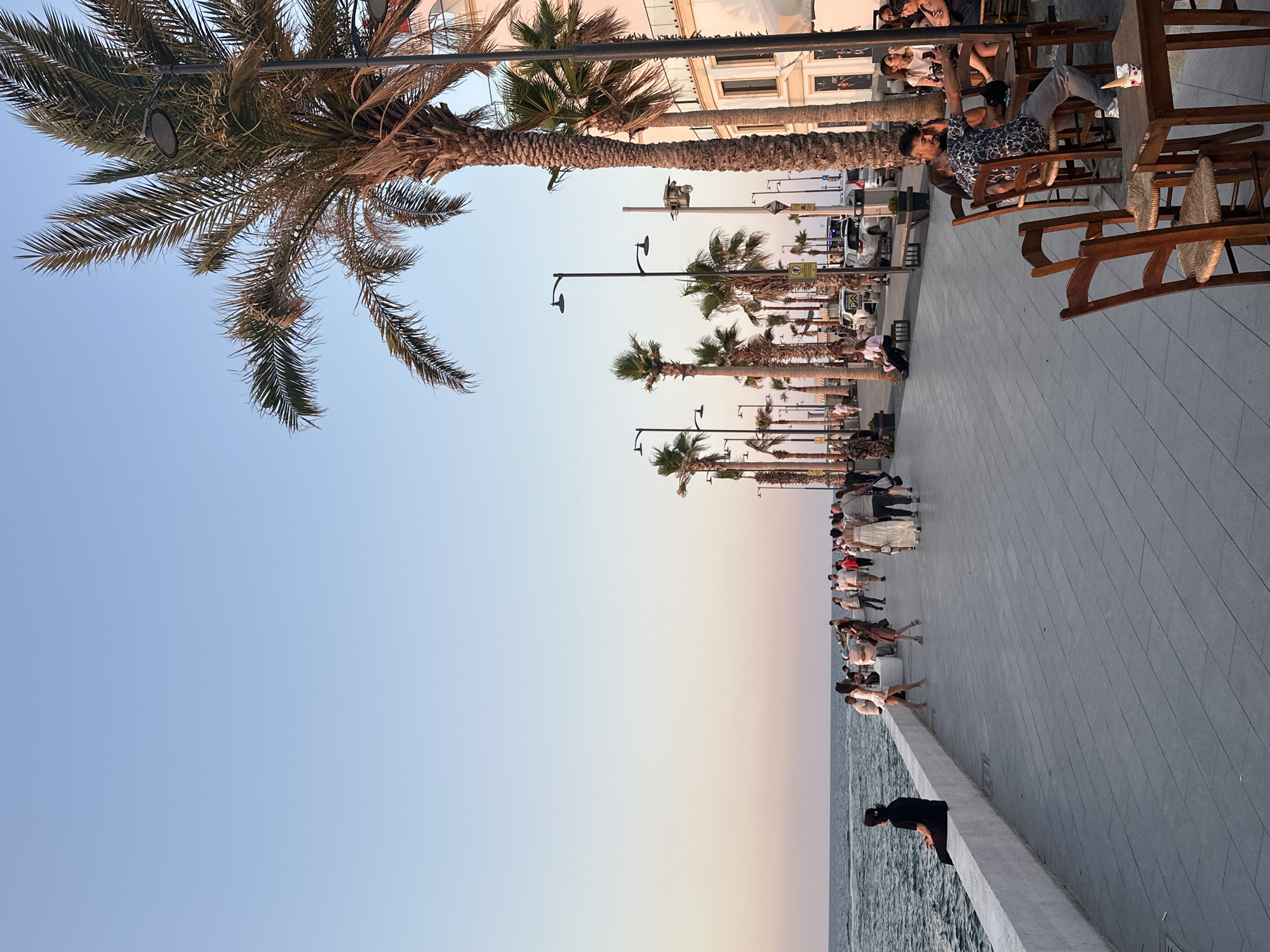 The image depicts a waterfront promenade in Kyrenia, Northern Cyprus, during what appears to be the late afternoon or early evening. The sky transitions from a pale blue at the top to soft hues of pink and orange near the horizon, suggesting a sunset. A paved walkway runs parallel to the sea, lined with palm trees and lampposts.
On the left side of the promenade, a white concrete barrier separates the walkway from the sea, which appears calm with gentle waves. A lone figure in a black t-shirt and dark pants is seated on the barrier, facing the water.
The promenade is populated by numerous people strolling and gathering. Some are walking in groups, while others are seated at tables outside a building that flanks the right side of the walkway. This building appears to be a multi-story structure with balconies and windows, exhibiting light-colored walls.
In the foreground on the right, several wooden tables and chairs are set up outside what might be a cafe or restaurant. A couple is seated at one of these tables; the man is holding a smartphone, appearing to take a selfie, while the woman leans towards him. An ice cream cup with a cone is visible on another table.
The palm trees are a prominent feature, with their fronds casting shadows on the walkway. In the background, several cars are parked along the street behind the promenade, and more buildings and palm trees are visible. The overall atmosphere is one of leisure and social activity.