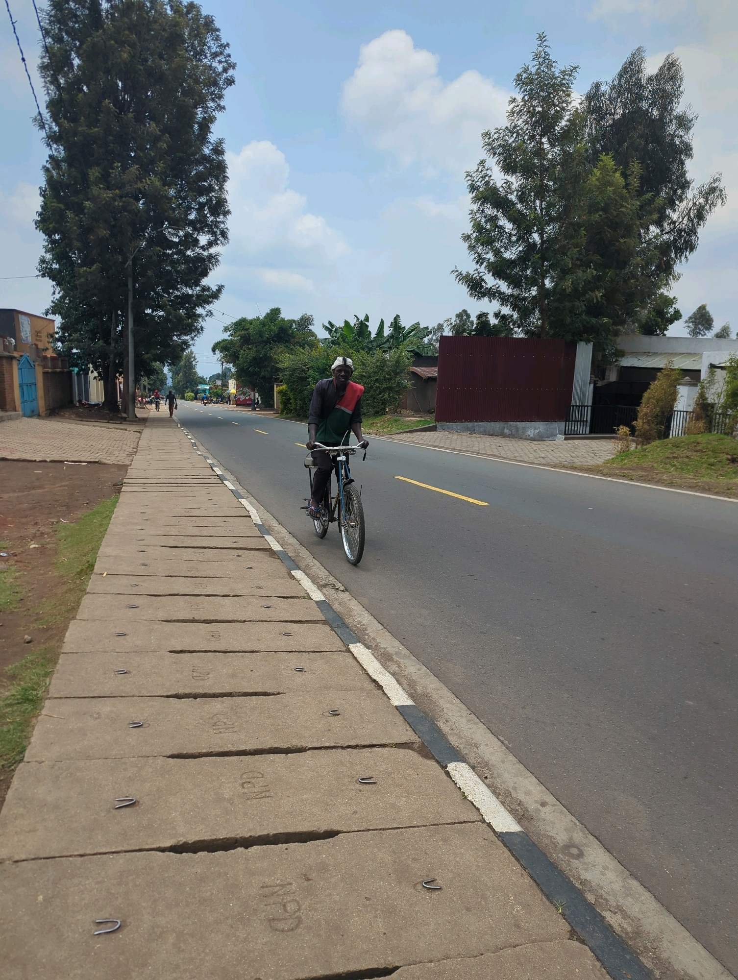 The image shows a man riding a bicycle on a paved road in Musanze District, Rwanda. The man is wearing a dark shirt and pants, and a white and red cloth is draped over his shoulders. He is smiling and looking towards the camera. The road is lined with a concrete sidewalk on the left side of the image. In the background, there are trees, buildings, and a few other people walking. The sky is partly cloudy. The scene appears to be a street in a residential or commercial area.