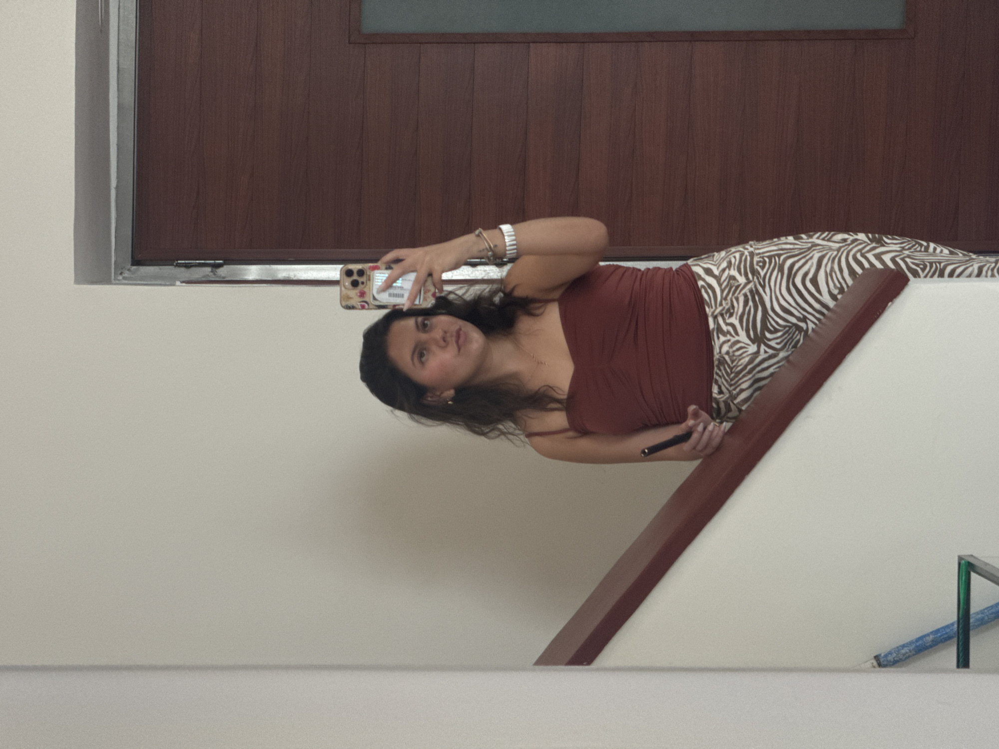 A woman is taking a selfie with a smartphone in a stairwell. She is wearing a brown top and zebra-striped pants. The background includes a wooden door with a grey frame and white walls. The location is Heroica Puebla de Zaragoza, Mexico.