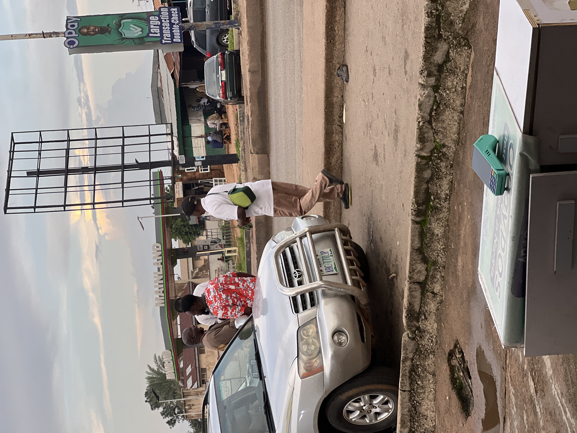 The image depicts a street scene in an urban environment, possibly in Nigeria, given the license plate format. In the foreground, a silver SUV is parked. Several individuals are gathered near the SUV, engaged in conversation. One man, wearing a red and white patterned shirt, is facing away from the camera. Another man in a brown garment is talking to him. To the right, a man in a white shirt and brown pants walks away from the camera, carrying a light green bag over his shoulder. In the background, a gas station with the "MAINLAND" signage is visible, along with various buildings and a large billboard structure. A dark sedan and a darker SUV are also present on the road. The ground is paved, with visible cracks and some debris. In the immediate foreground, a green Point of Sale (POS) terminal rests on a counter. The overall lighting suggests it is either early morning or late afternoon, with the sky showing some cloud cover.
