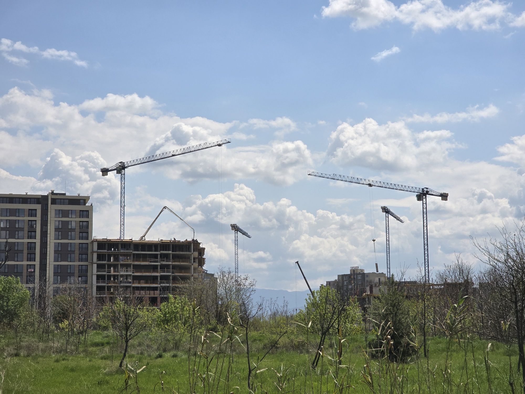 The image depicts a construction site in Unknown, Unknown. Several cranes are visible, along with partially constructed buildings. The foreground is characterized by green grass and vegetation. The sky is blue with scattered clouds.