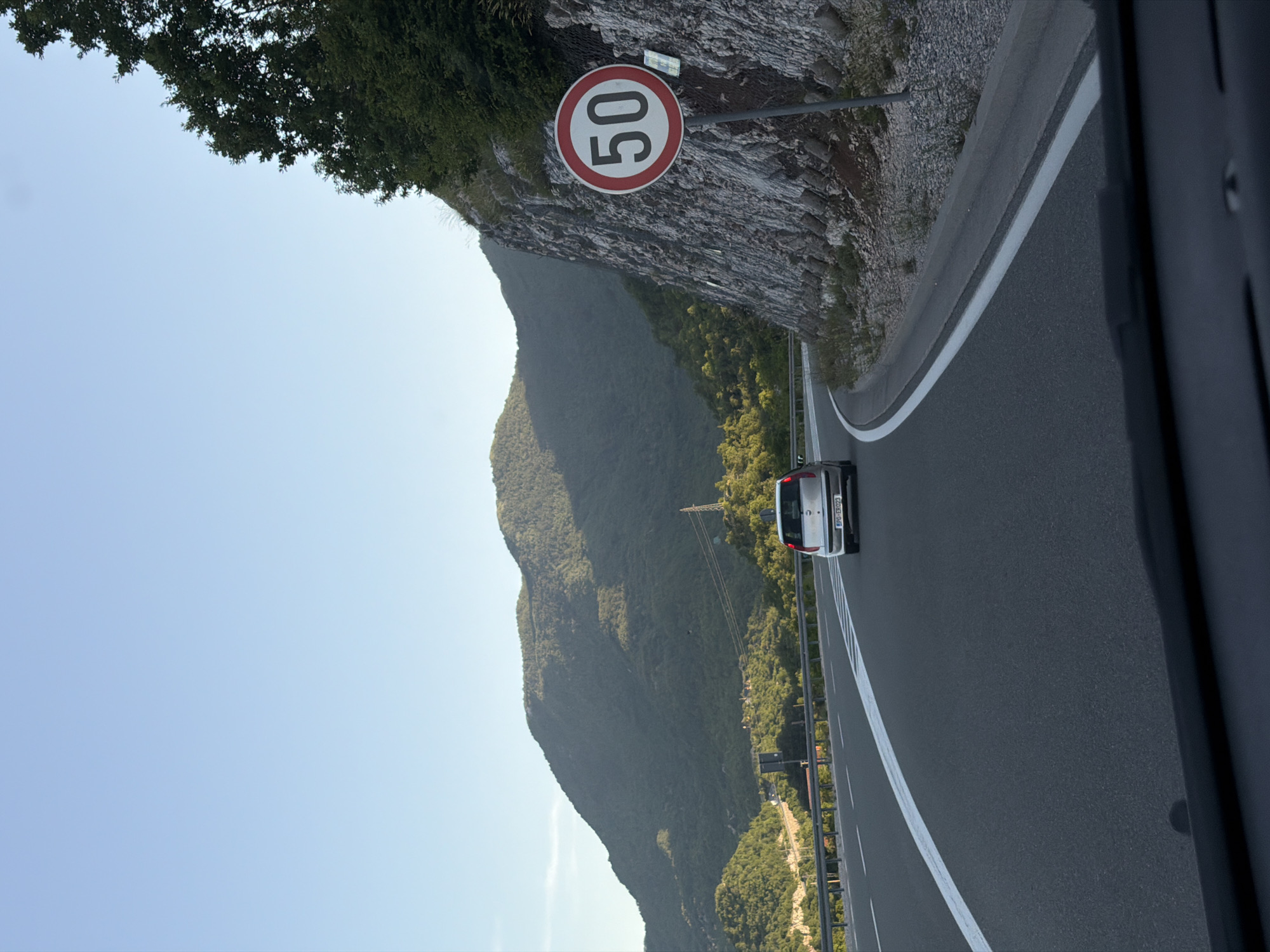 A white car is driving on a highway with a speed limit of 50 km/h. The highway is bordered by a rocky cliff face covered in trees on the right, and a forested mountain range on the left. The sky is clear and blue. The car's license plate reads "PG-EK023". The scene appears to be in Vrela, Montenegro.
