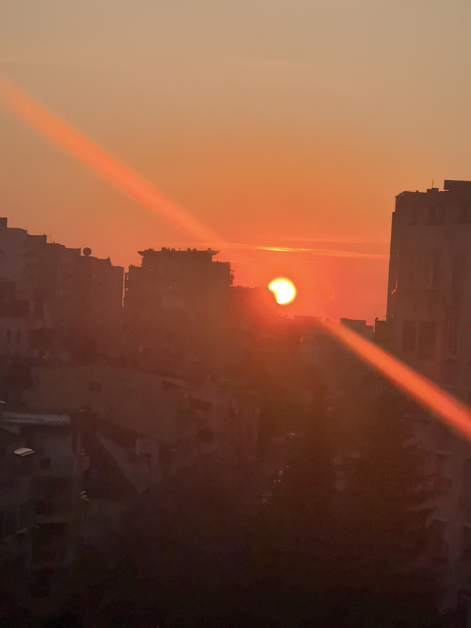 The image captures a striking partial solar eclipse, likely during sunrise or sunset, over an urban landscape in Stara Zagora, Bulgaria. The sky displays a vibrant gradient, transitioning from a soft peach hue at the top to intense orange and fiery red tones near the horizon.
The central focus is the sun, visible as a brilliant, crescent-shaped light source, indicative of a partial eclipse. A fainter, ghost-like crescent image of the sun is also discernible slightly to its right, likely an internal lens reflection. A prominent, diagonal orange-red lens flare streaks across the frame, originating from the sun and extending both upwards and downwards, adding to the dramatic effect.
Silhouetted against the bright sky are multiple multi-story buildings, typical of an urban environment. These structures, appearing dark brown or black, frame the scene on the left, right, and partially across the horizon line, with some rooftops revealing antennas or architectural details. The lower foreground is very dark, showing indistinct shapes of further buildings or ground features, heavily underexposed by the bright sky.
The overall scene conveys a clear, though potentially hazy, atmosphere, allowing the celestial event and the warm colors of the sky to dominate. There is no visible text in the image.