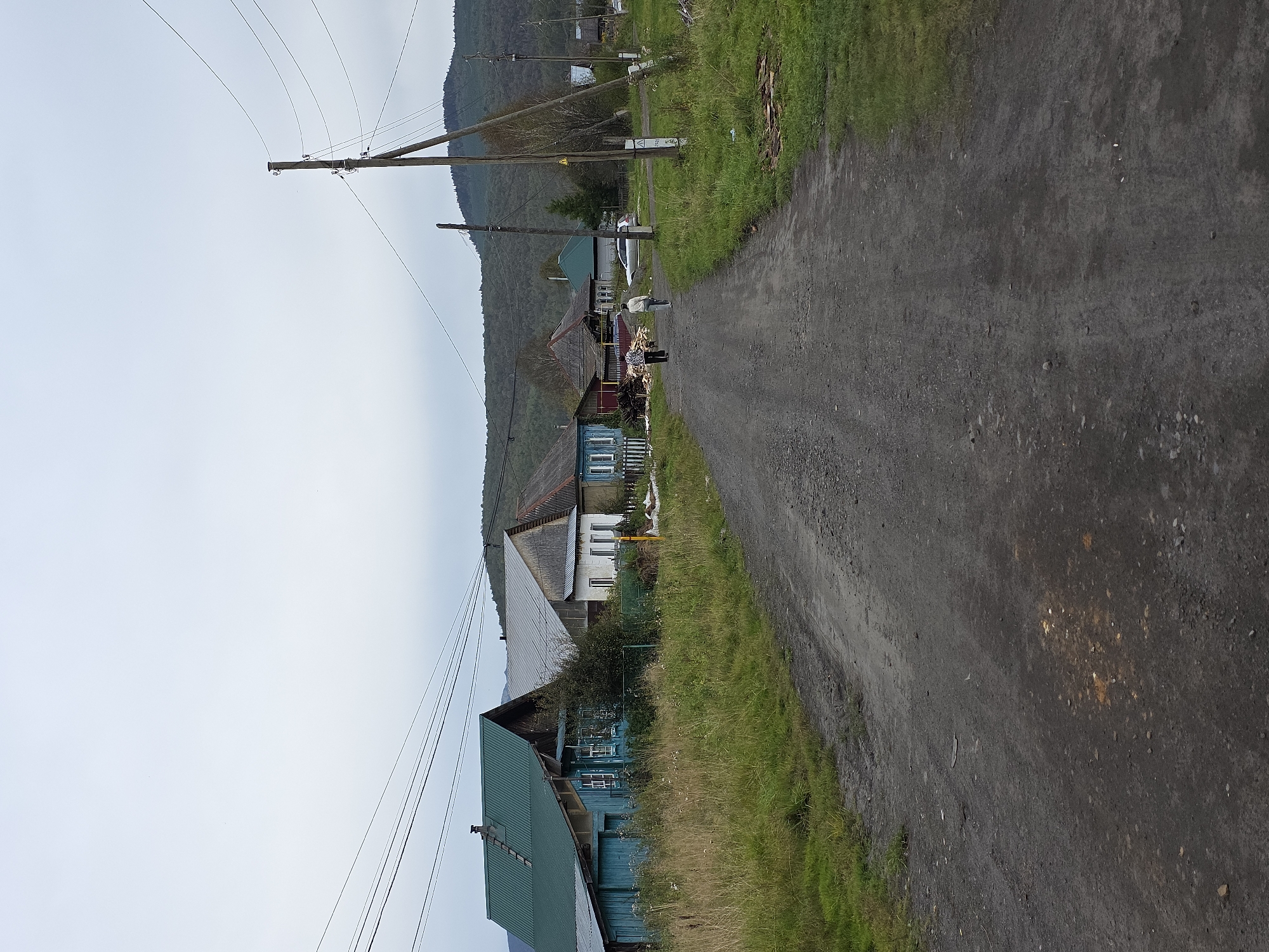 In Magnitka, Russia, a dirt road extends through a rural village under an overcast sky. The road is flanked by a ditch and overgrown grass. On the left, a cluster of traditional wooden houses with steeply pitched roofs in green and gray dominate the scene. The houses feature blue accents and visible windows. Further down the road, two individuals, one in a dark shirt and pants and the other in a light shirt and pants, walk away from the camera. On the right side of the road, a wooden utility pole with wires runs overhead, and a line of trees and rolling hills forms the backdrop. Additional houses are visible in the distance.