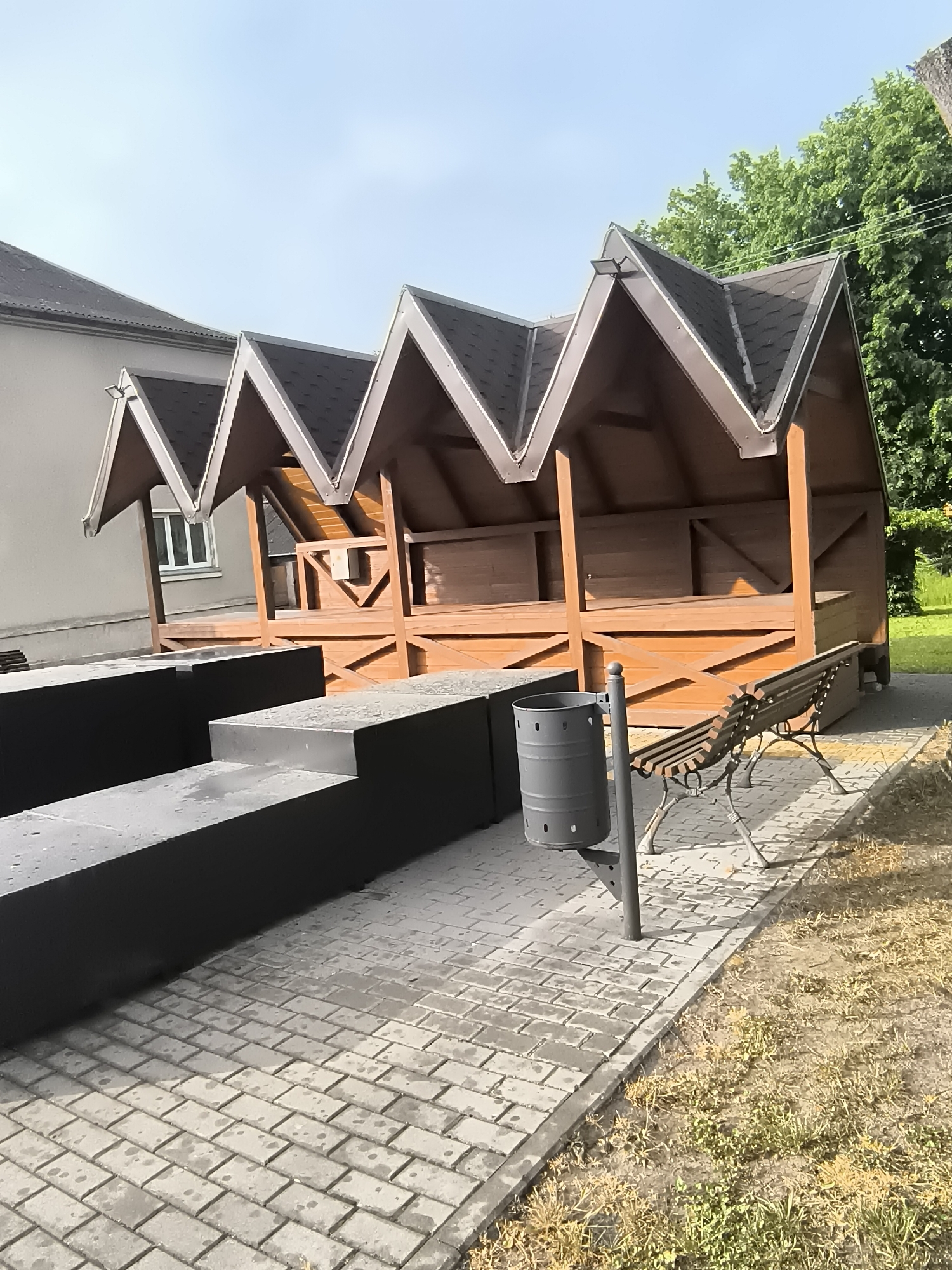This outdoor scene in Priekulė, Lithuania, captures a quiet public area featuring a row of three distinct wooden shelters or gazebos. These structures are characterized by their striking zig-zagging dark brown roofs, which are shingled, and light brown wooden frames. Each gazebo is open-sided, with wooden railings and benches within, suggesting a place for relaxation or gathering.
In the foreground, a series of dark grey, rectangular planters or low walls are arranged in a tiered fashion, running parallel to the gazebos. Adjacent to these, a grey paved walkway made of interlocking bricks stretches across the midground. To the right of the paving, a dark grey cylindrical trash can is mounted on a pole. Further right, a traditional park bench with a slatted wooden seat and back, supported by ornate dark metal legs, is positioned on the paving.
Behind the gazebos, a light-colored, possibly stucco, building with a dark roof and a single visible window is present. Green trees and foliage are visible in the background, especially to the right, indicating a natural green space nearby. The sky above is clear and light blue, suggesting a bright and sunny day, likely morning or early afternoon, as evidenced by the distinct shadows cast on the paving and structures. No people or specific activities are visible, lending a peaceful and deserted atmosphere to the scene. There is no visible text in the image.