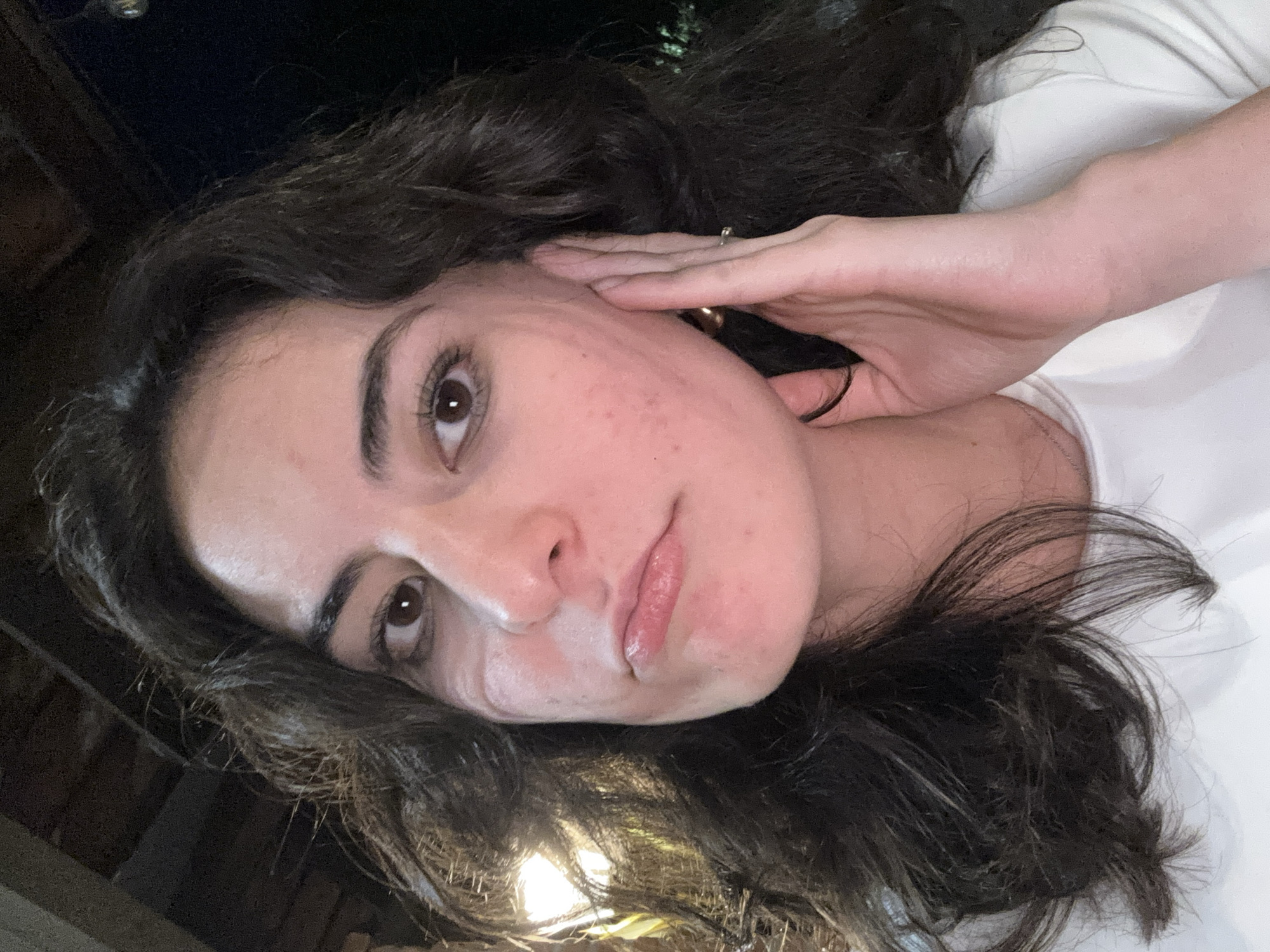 A young woman with dark, wavy hair and brown eyes is posing for a selfie. She is wearing a white shirt. Her left hand is raised, partially covering her cheek and ear. A ring is visible on her finger. The background is dark and indistinct. The location is unknown.