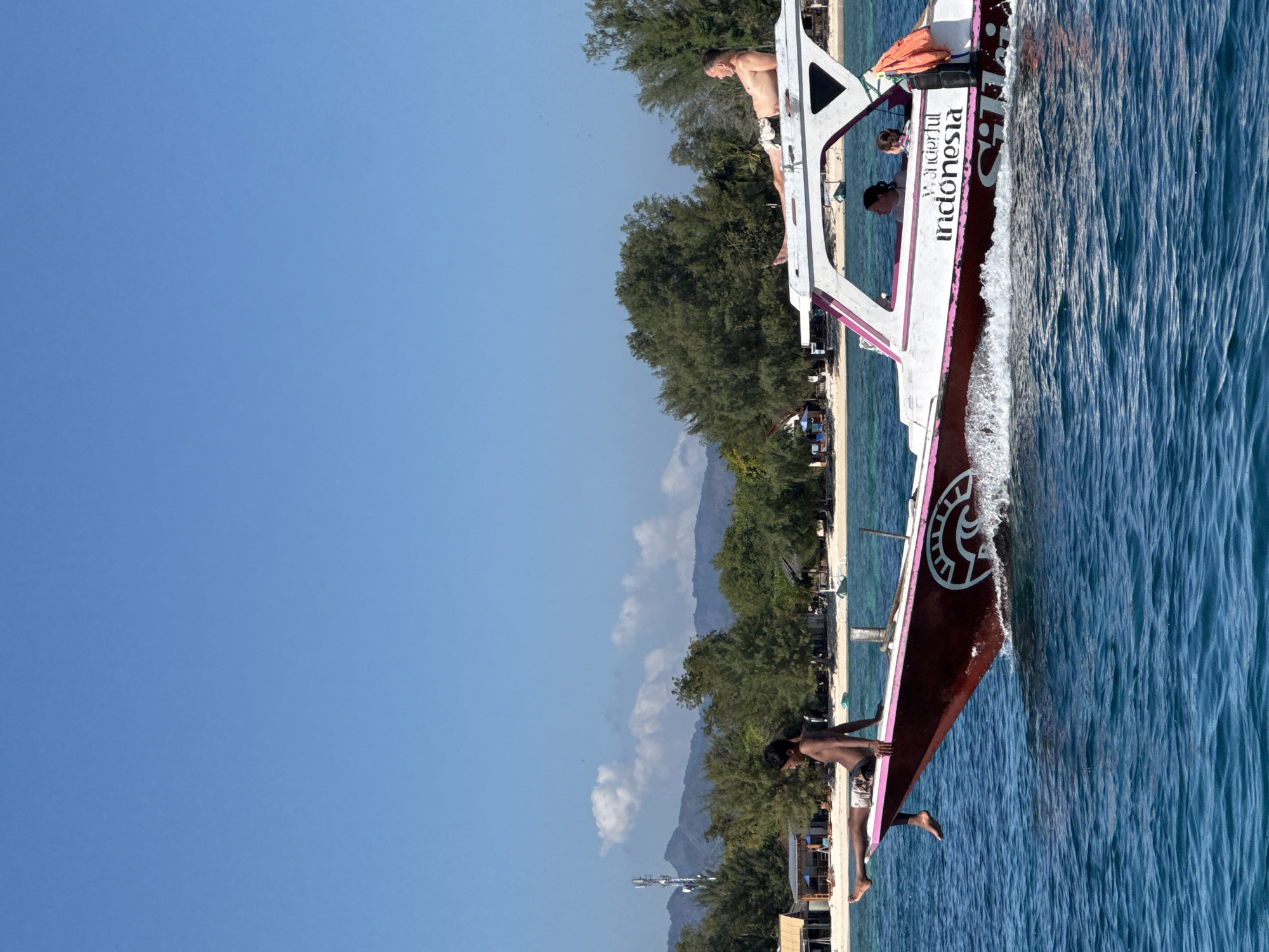 The image shows a motorboat with a maroon hull and white cabin, speeding through blue water under a clear sky. The boat has the text "Wonderful Indonesia" printed on its side. There are people on the boat: a young person sitting on the front edge of the boat with their legs dangling over the water, and an adult lying down on top of the cabin. Inside the cabin, two women are visible, one of whom appears to be operating the boat. In the background, a tropical island with lush green trees, white sand beaches, and some small buildings is visible. Mountains can be seen further inland, with a tall communication tower on one of them. The water is choppy, indicating the boat's speed.