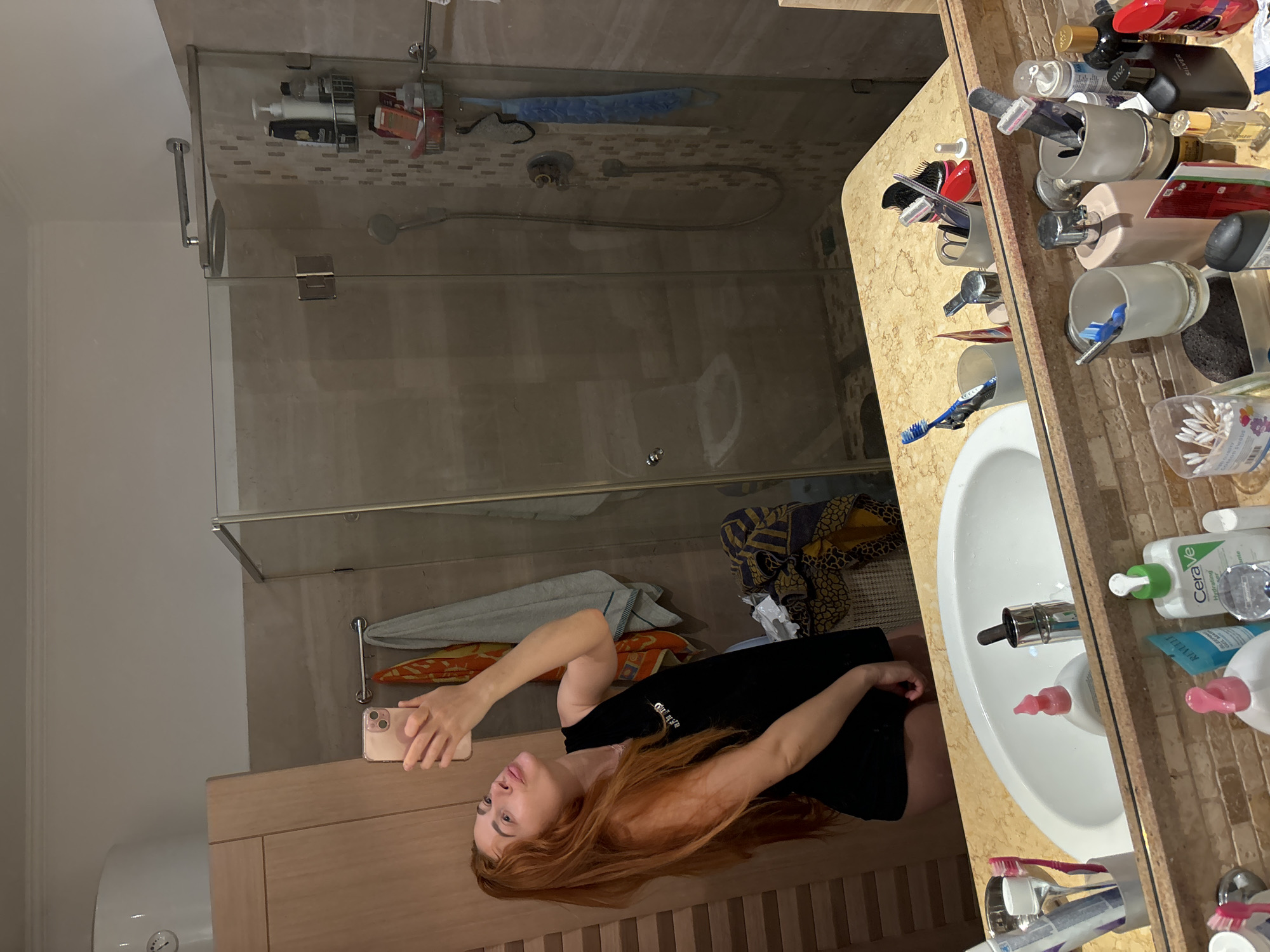 An indoor full-shot depicts a young woman with long, red hair taking a mirror selfie in a well-lit bathroom, likely in Budva, Montenegro. The original image is rotated 90 degrees counter-clockwise.
The woman, wearing a black top, holds a pink smartphone with her right hand, capturing her reflection in a large mirror. Her gaze is directed towards the phone. The mirror extends over a beige, granite-patterned vanity counter.
The vanity features a white ceramic sink with a modern metal faucet and a pink liquid soap dispenser. The counter is extensively cluttered with numerous personal care items and toiletries, including various bottles, combs, razors, and toothbrushes stored in glass holders. Notable items include a CeraVe Hydrating Cleanser bottle, a bottle marked "silver," and other hair and skin products. A brown pumice stone is also visible near the sink.
Behind the woman's reflection, a shower enclosure with clear glass partitions is visible. The shower walls are tiled with small, light brown rectangular tiles. Inside the shower, a metal caddy holds additional toiletries, and a shower head with a hose is present. Towels in various colors (grey, orange, blue) are draped over a rack within the shower area.
To the left of the vanity, a wooden cabinet provides storage. A white water heater or boiler with a gauge is partially visible on the bottom left. The overall setting is a functional and lived-in bathroom.