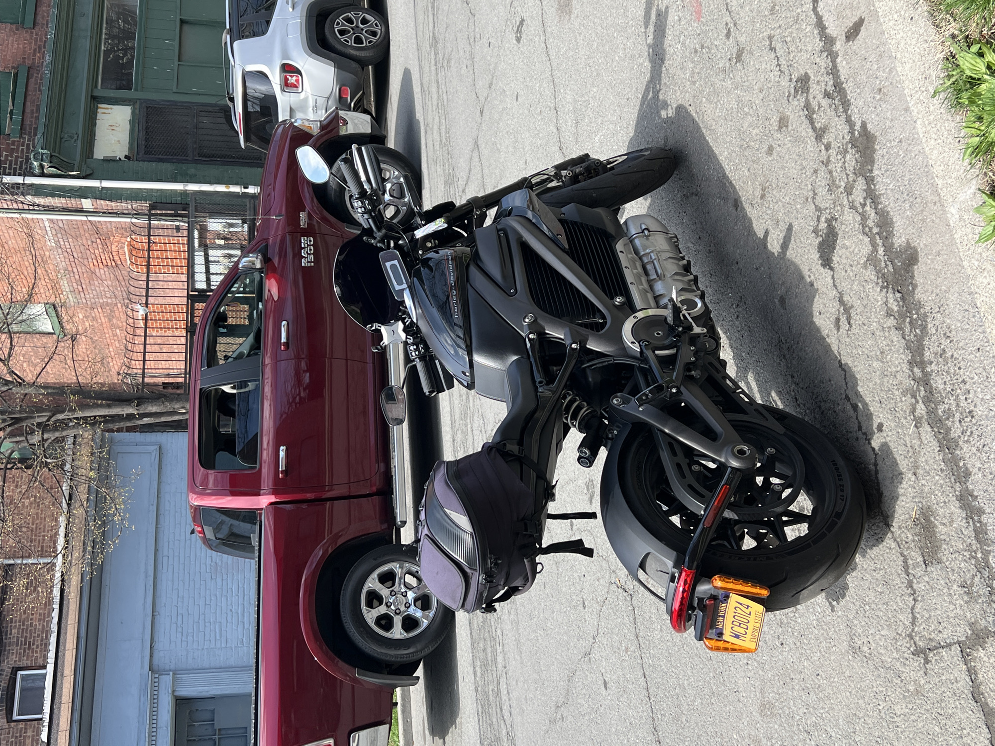 The image shows a black Harley-Davidson motorcycle parked on a city street in what appears to be the City of Newburgh, United States of America. The motorcycle is positioned at an angle in relation to the curb, and has a license plate visible with the characters "MCB0124". A burgundy Ram 1500 pickup truck is parked directly behind the motorcycle, and a silver Jeep is visible parked behind the truck. The background includes building facades, a metal fence, and trees with sparse foliage, suggesting a residential or mixed-use urban environment. The time of day appears to be daylight, with the sun casting shadows.