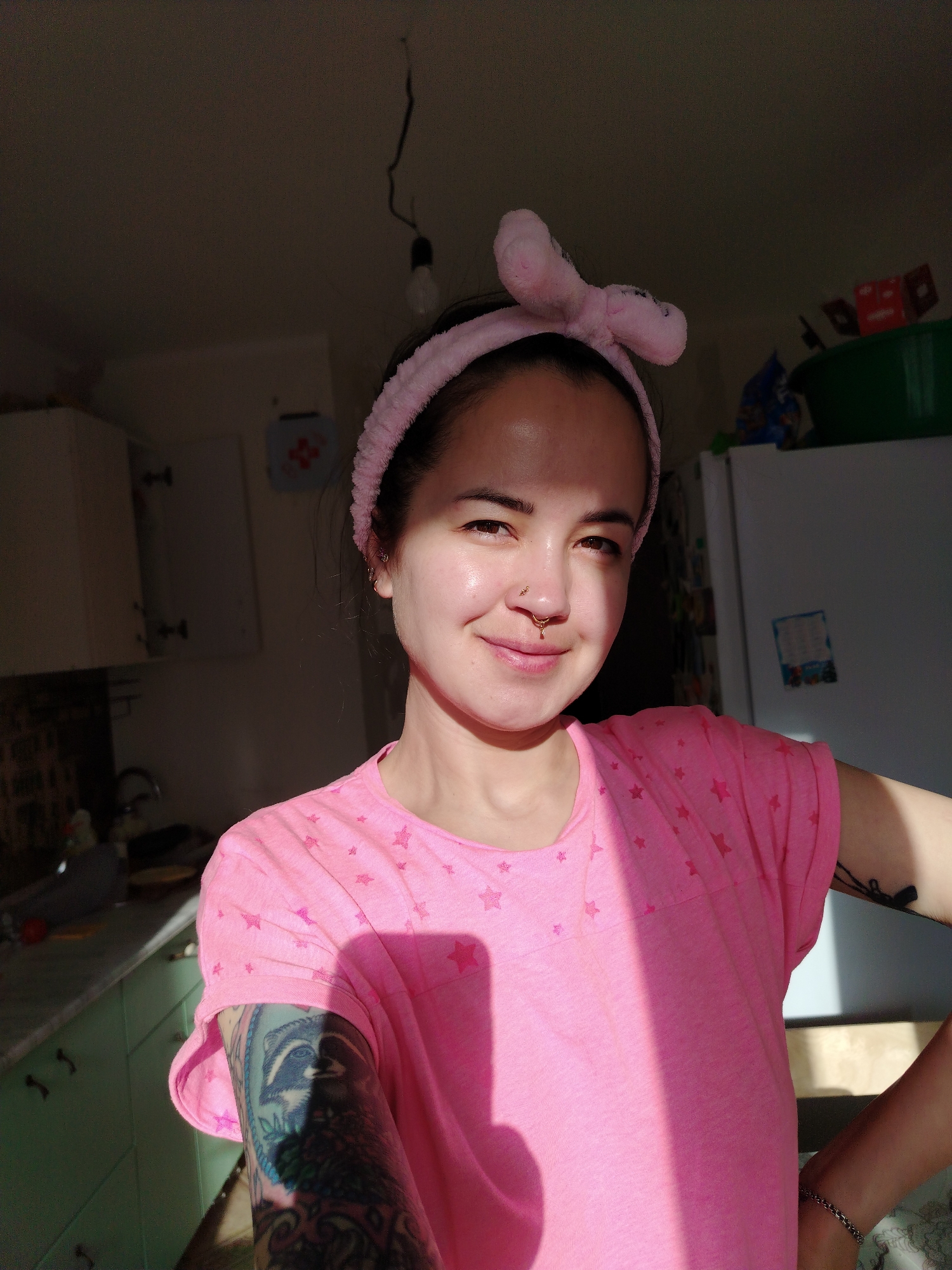 A young woman poses indoors in a brightly lit room, likely a kitchen, given the presence of cabinets, a sink, and a refrigerator. She is wearing a pink t-shirt with star patterns and a pink headband with a bow. She has a nose piercing and tattoos on her left arm. The lighting casts shadows across her face and torso. A lightbulb hangs from the ceiling. The location is stated as Noviy, Russia.