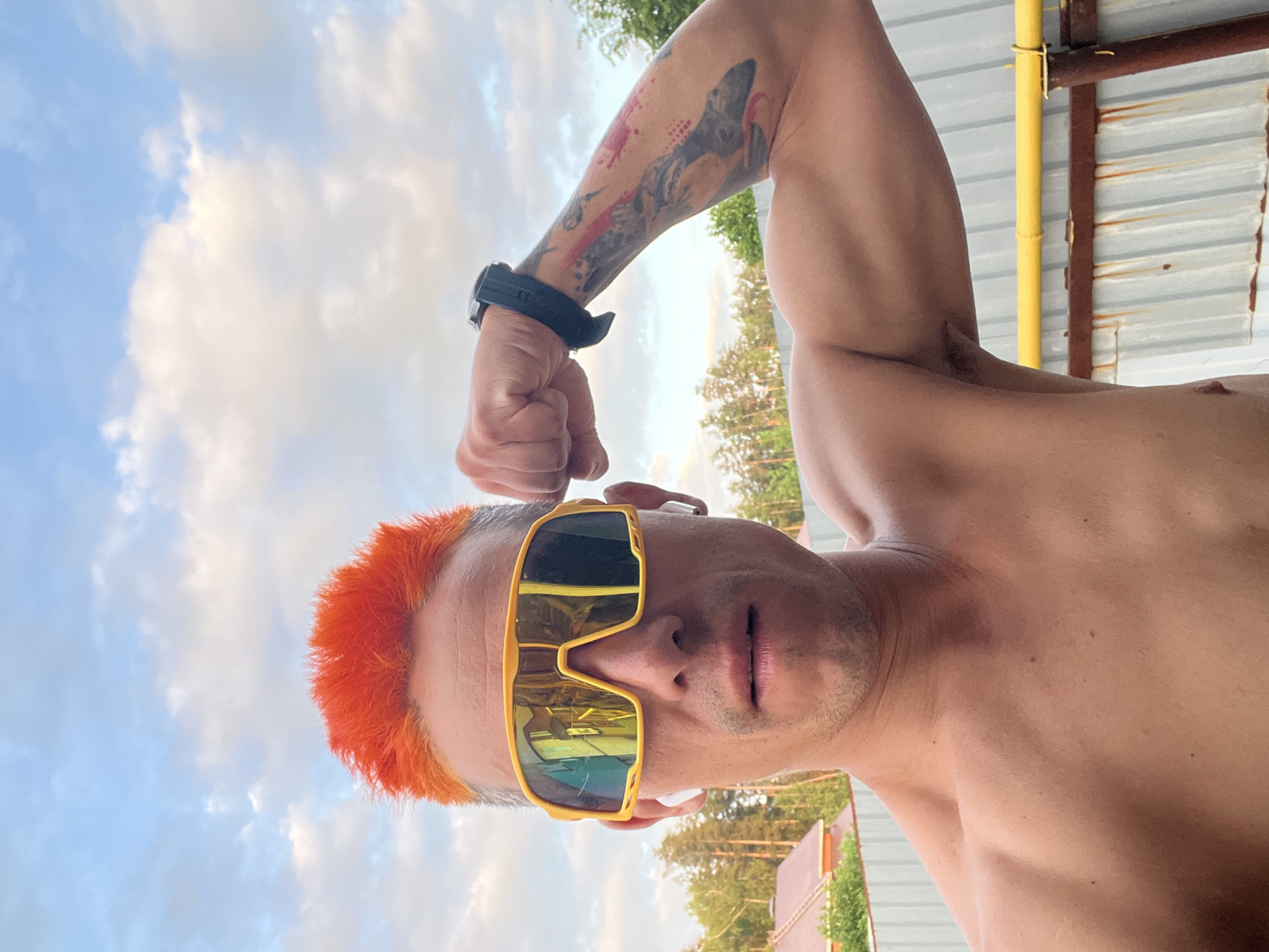 A low-angle, slightly overexposed, waist-up shot depicts a shirtless man with orange hair, wearing gold-framed sunglasses and a black wrist-worn device. He is flexing his left arm, which has a tattoo. The background includes a partly cloudy sky, trees, and a metallic structure with yellow supports. The location is likely Chaikovsky, Russia, based on the presence of the metallic structure, which is typical in the area.