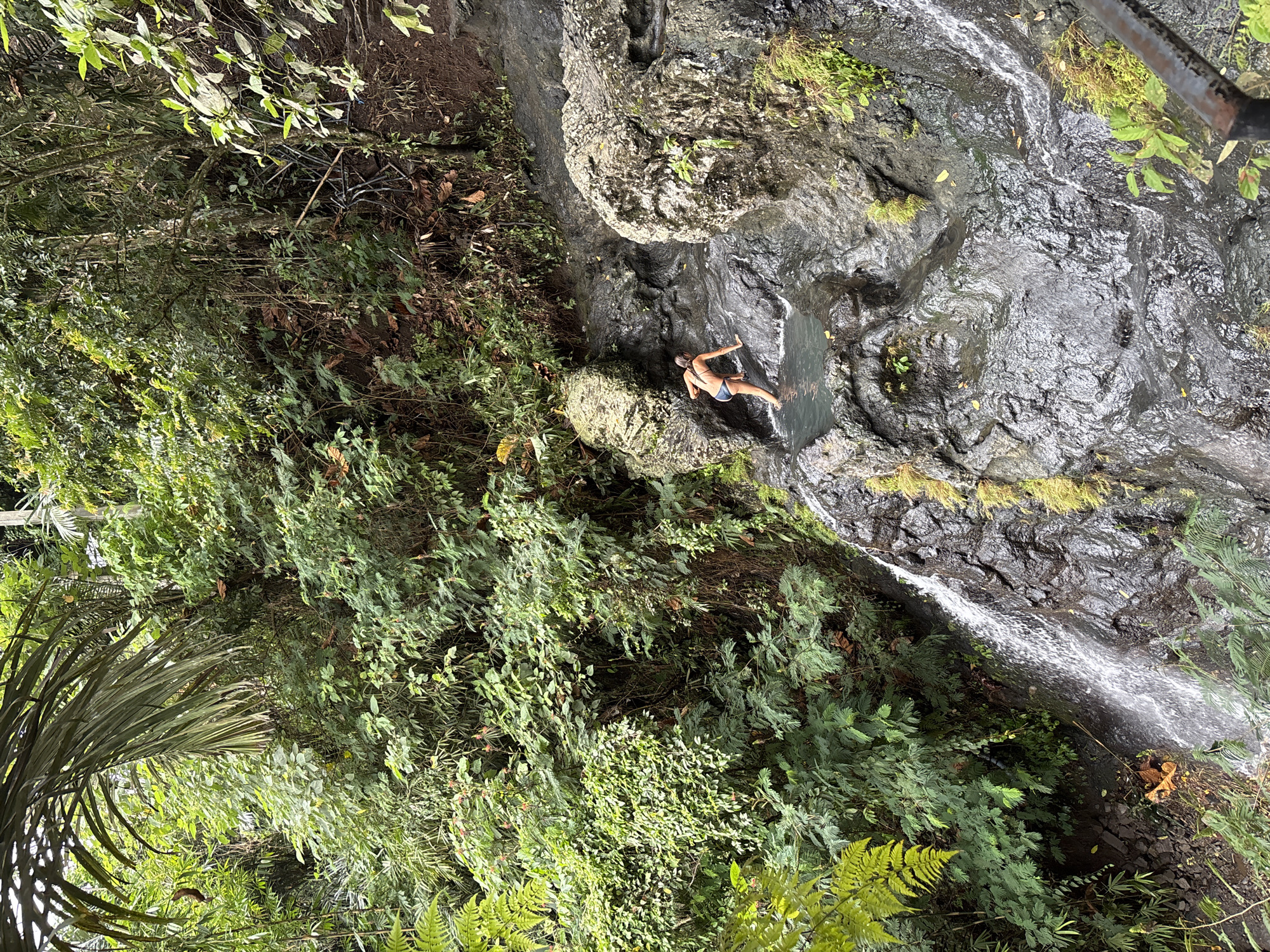A person in a bikini wades through a shallow pool of water at the base of a waterfall in a lush, green jungle. The waterfall cascades down a rocky incline covered in moss and vegetation. The scene is set in Sidemen, Indonesia, a region known for its natural beauty and rice paddies. The water is clear and reflects the surrounding greenery. The person appears to be enjoying the refreshing water, with their back to the camera. The overall atmosphere is serene and tranquil, with the sounds of nature dominating the scene.
