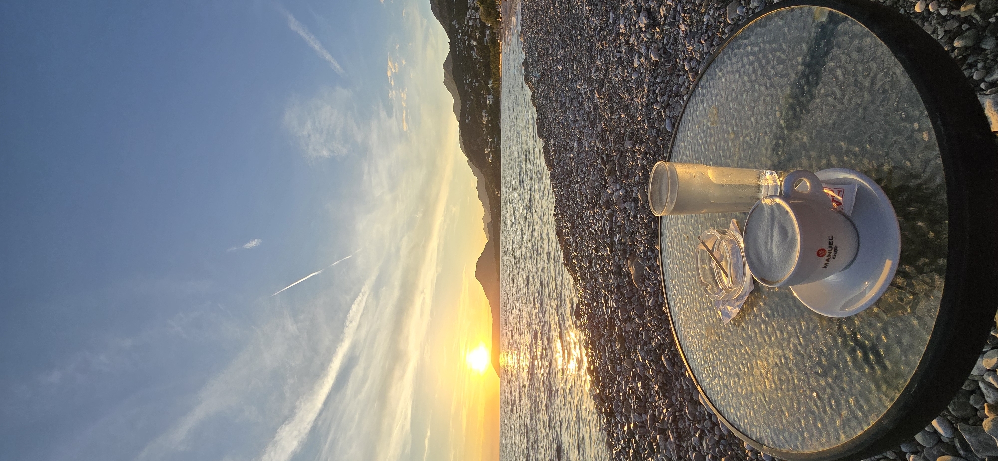 This media file presents a tranquil scene of either sunrise or sunset over a pebble beach and the sea, likely in Bar, Montenegro. The sun, a brilliant golden orb, is positioned low on the horizon, casting warm light across the water, creating a shimmering path of reflections.
In the foreground, on the right side, there's a round table with a textured glass top. On the table, a white coffee cup with a saucer and a clear glass are visible. The coffee cup distinctly displays the brand "MANUEL Caffè" in red lettering with a logo. Next to these items, a small, clear glass ashtray holds a stirring stick.
The middle ground features a calm, gently rippling sea and a wide expanse of pebble beach that extends along the shore. The background reveals a range of mountains, their silhouettes softened by the golden hour light, with several white buildings discernible on their slopes, indicative of a coastal settlement. The sky is a clear blue, graduating to warmer tones near the horizon, with delicate, wispy clouds and a few visible airplane contrails. The overall mood conveyed is one of peaceful relaxation, capturing a beautiful moment by the sea.