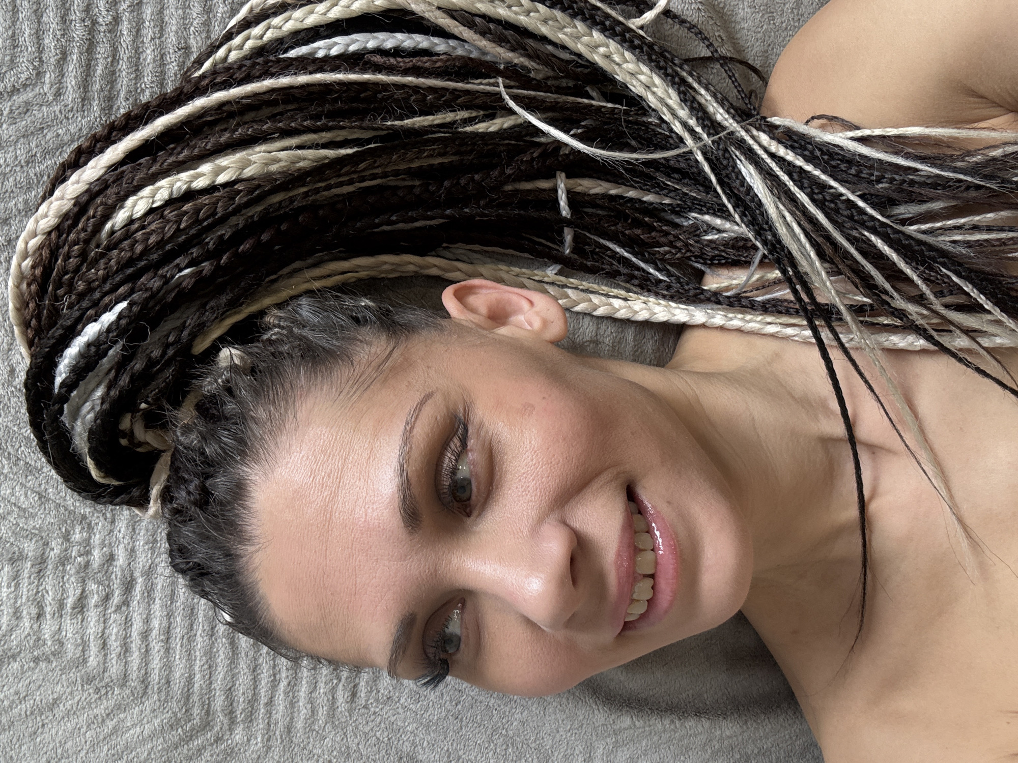 A close-up, angled, high-angle shot presents a woman with braided hair lying on what appears to be a gray fabric surface. The braids are a mix of dark brown, black, and light blonde or off-white colors, cascading around the left side of her face and upper chest. Her head is slightly tilted, and she is smiling, revealing her teeth. Her eyes are open and appear to be light-colored. The lighting is soft and diffused, illuminating her face and hair. The texture of the gray fabric in the background indicates that it could be a towel or a blanket.