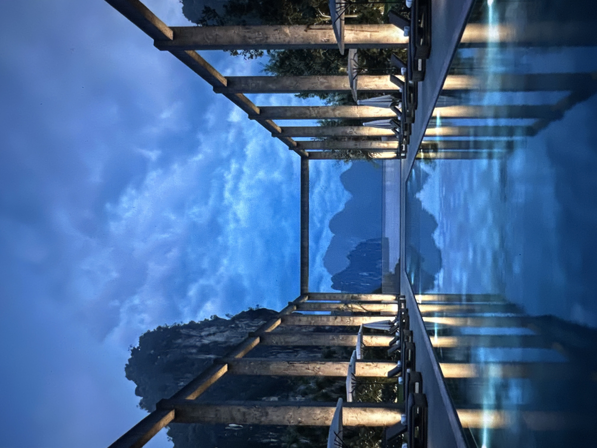 An outdoor infinity swimming pool is illuminated by overhead lights and reflected in the dark water, creating a serene atmosphere. The pool is flanked by rows of lounge chairs and umbrellas, suggesting a resort or hotel setting. In the background, majestic mountains rise under a twilight sky, their peaks shrouded in mist. The scene is framed by a modern architectural structure with concrete pillars and beams, adding a sense of grandeur and sophistication. The overall ambiance is one of tranquil luxury, with a harmonious blend of natural beauty and man-made design.