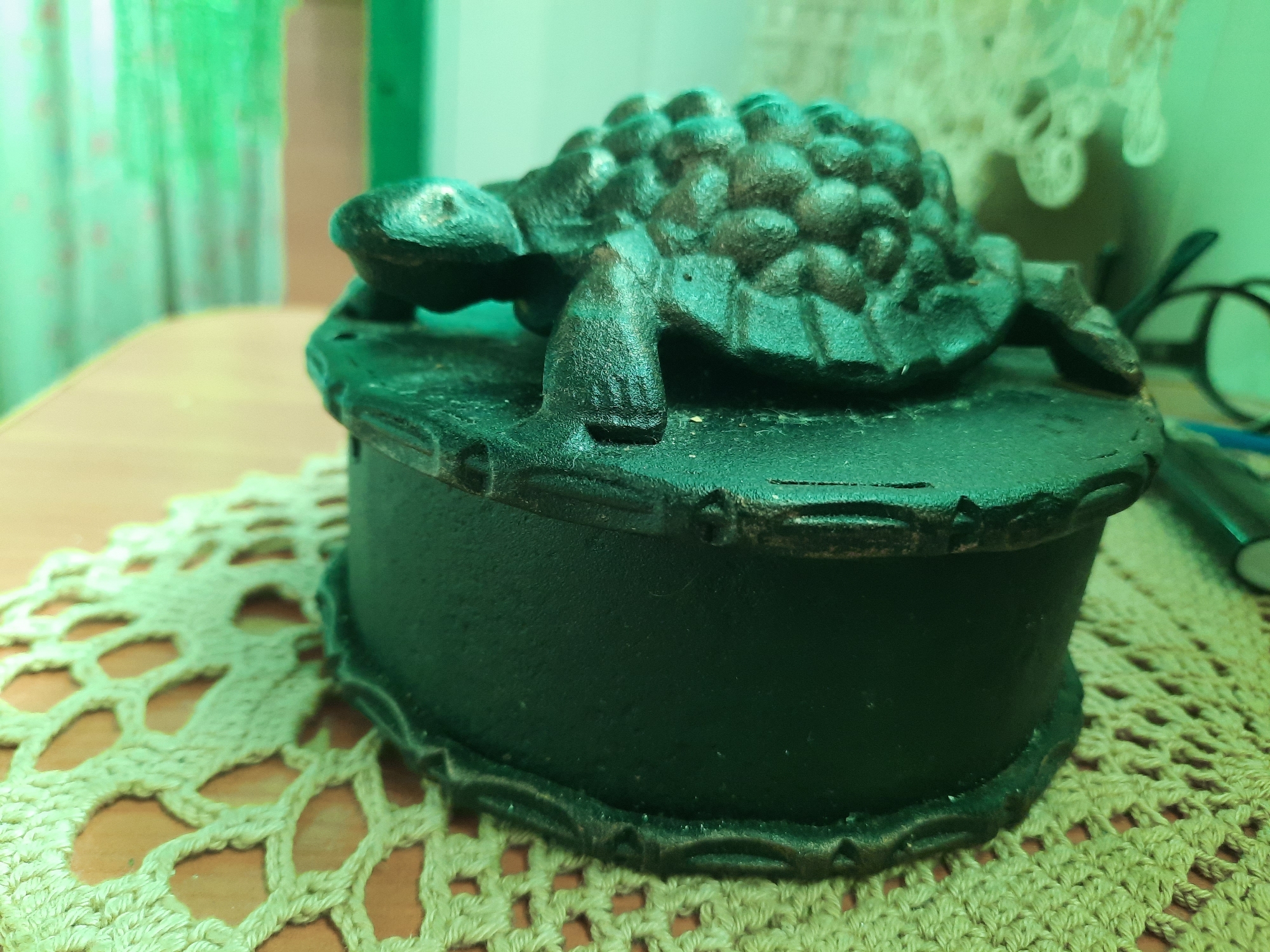 A dark, ornate, possibly cast-iron box sits on a crocheted doily. A decorative turtle figurine is attached to the lid of the box, its shell textured and its legs and head detailed. The box itself is dark green and hexagonal. The doily is a pale yellow-green color with an intricate openwork pattern. The background is blurred and appears to be an indoor setting, with hints of green curtains and a wooden table surface. The lighting casts a green hue over the entire scene. There are no people visible in the image. The scene is a still life arrangement of decorative objects.