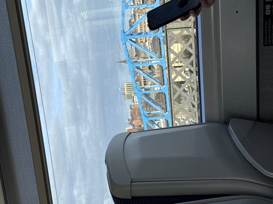 From a train window, the iconic blue framework of Tower Bridge dominates the view. In the background, a cityscape emerges, punctuated by a church spire. A hand holds a phone, partially obscuring the scene. The interior of the train is visible, featuring a light grey seat with a fold-down tray table. A sign indicating power outlets is on the wall.