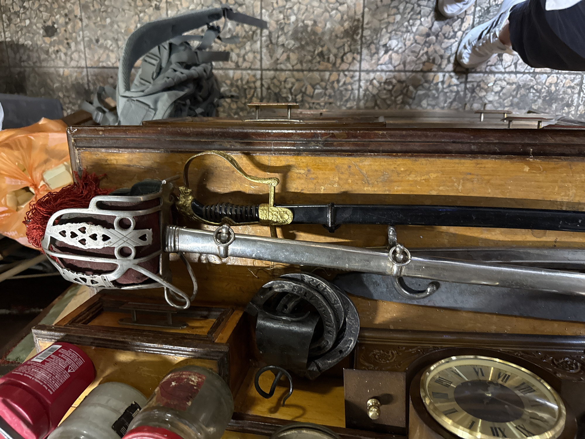 The image depicts a close-up, overhead view of several objects placed on a wooden surface. In the upper left, there are two glass jars, one with a red lid and a red plastic container. To the right of these, a decorative helmet with metallic accents and red fringe is visible. Two swords, one with a silver blade and ornate hilt, and another with a black scabbard and a gold-colored hilt, are positioned vertically. Below the helmet, a stack of metal objects, possibly horseshoes, are piled. To the left, a circular clock face with Roman numerals is mounted on the wooden surface. The background shows a tiled floor and the leg of a person wearing white sneakers and dark pants. The scene appears to be an indoor setting, possibly a room or a display area. No specific location in Perm, Russia, can be identified from the provided image.