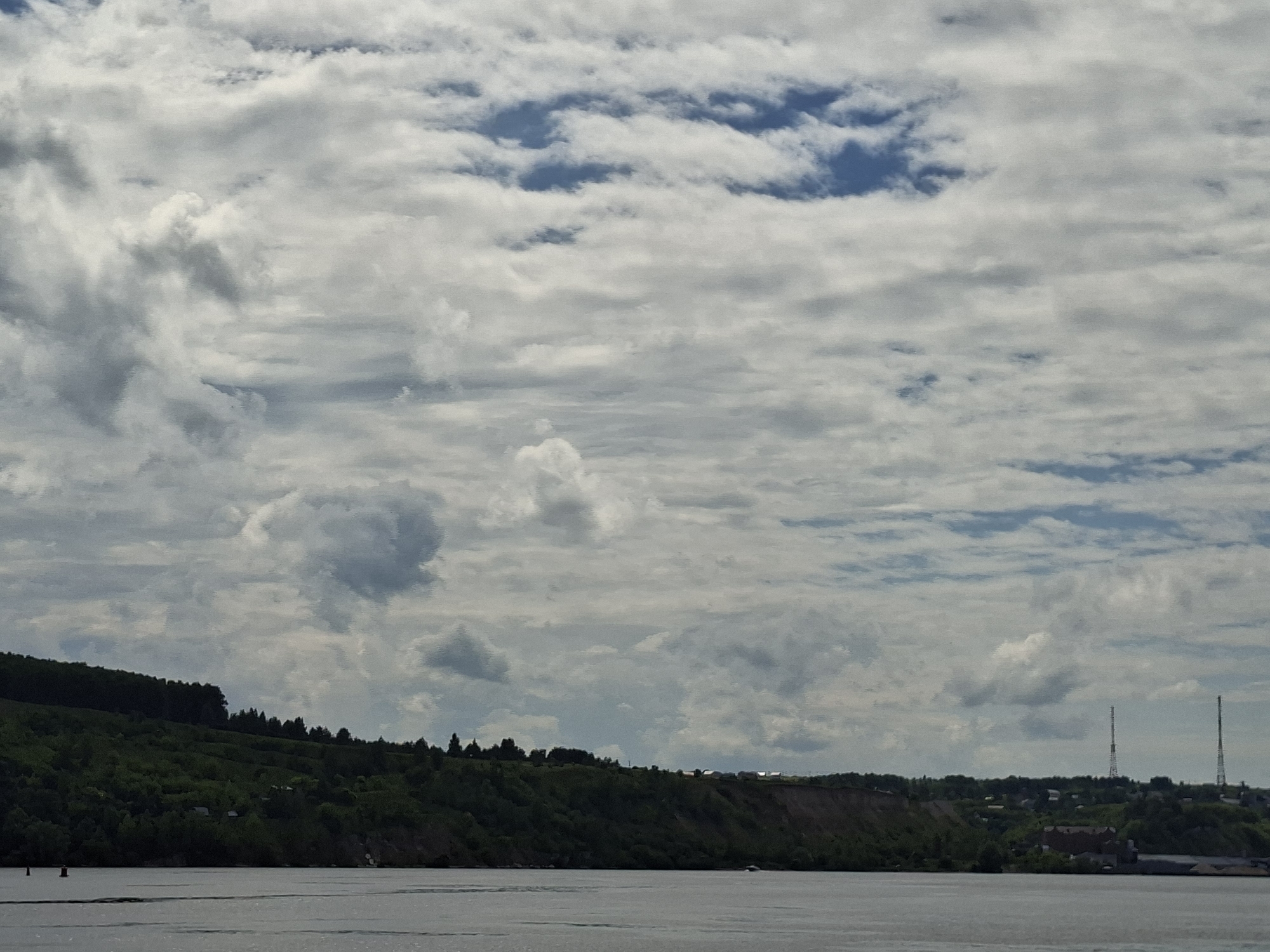 The image shows a panoramic view of the Volga River in Kazan, Russia, under a cloudy sky. The foreground features the calm, gray waters of the river with slight ripples. The middle ground displays a steep, tree-covered embankment on the left, transitioning to a more residential area with buildings and vegetation along the ridge. Two tall, slender radio towers are visible in the distance on the right side of the embankment, marking the city's skyline. The sky is filled with a mixture of white and gray clouds, with patches of blue sky showing through, indicating a dynamic weather condition. There are no people or active boats visible in the scene. The overall impression is one of a serene natural landscape meeting urban development.
