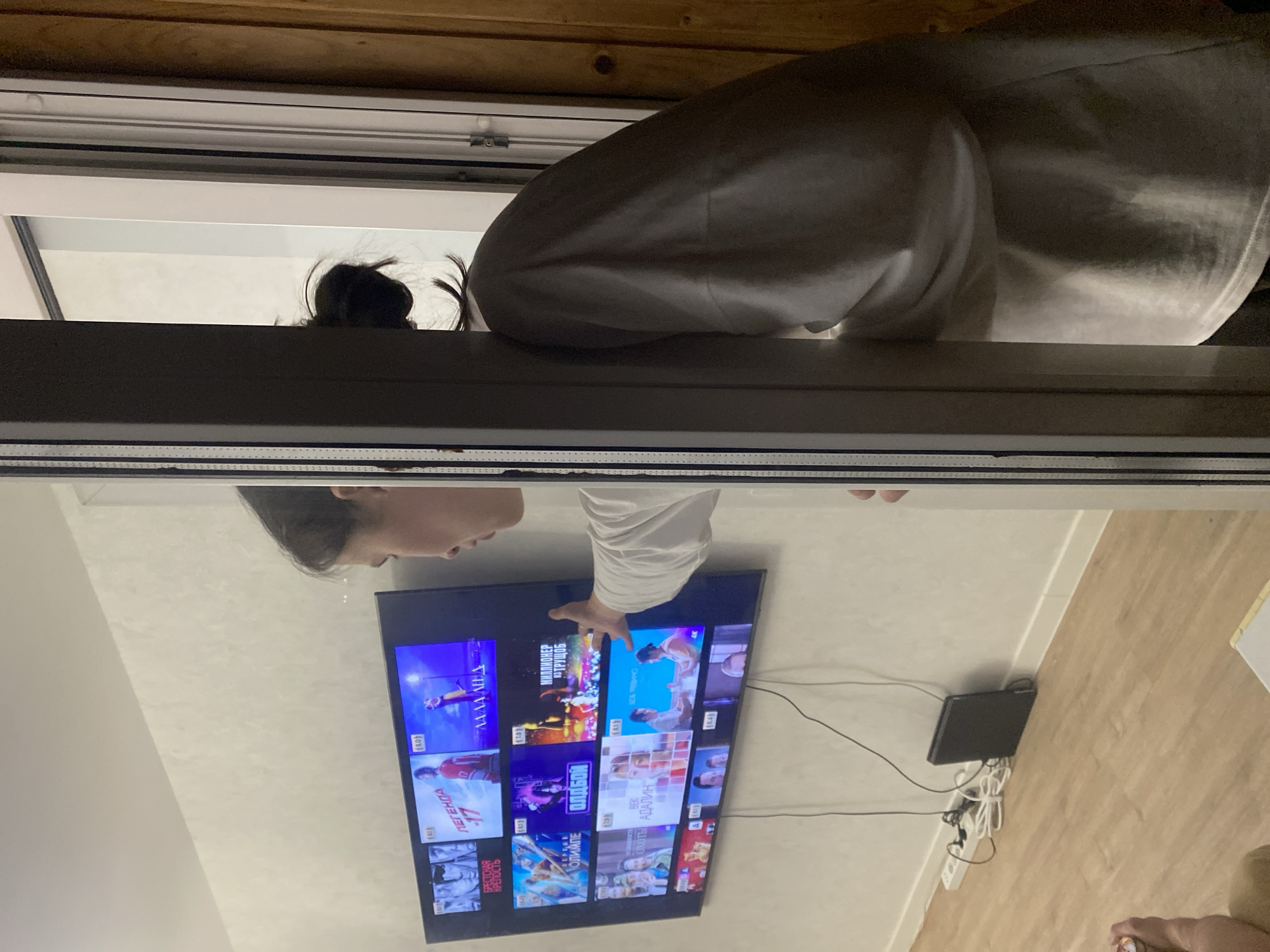 The image depicts a person with dark hair tied in a bun, wearing a light-colored sweatshirt, looking at a large television screen. The television displays a grid of movie posters or thumbnails, many with Russian titles and ratings. The person is reaching out towards the screen with their right hand, as if selecting something. The scene appears to be indoors, likely a living room, with light-colored walls and a wooden floor. In the background, a power strip and some cables are visible, along with a black electronic device. The person is partially framed by a doorway or window frame, with a wooden-paneled wall visible on the right side. The overall lighting suggests it might be evening or indoors with artificial lighting. The image does not contain any visual information that would allow for the specific identification of the city as Perm, Russia.