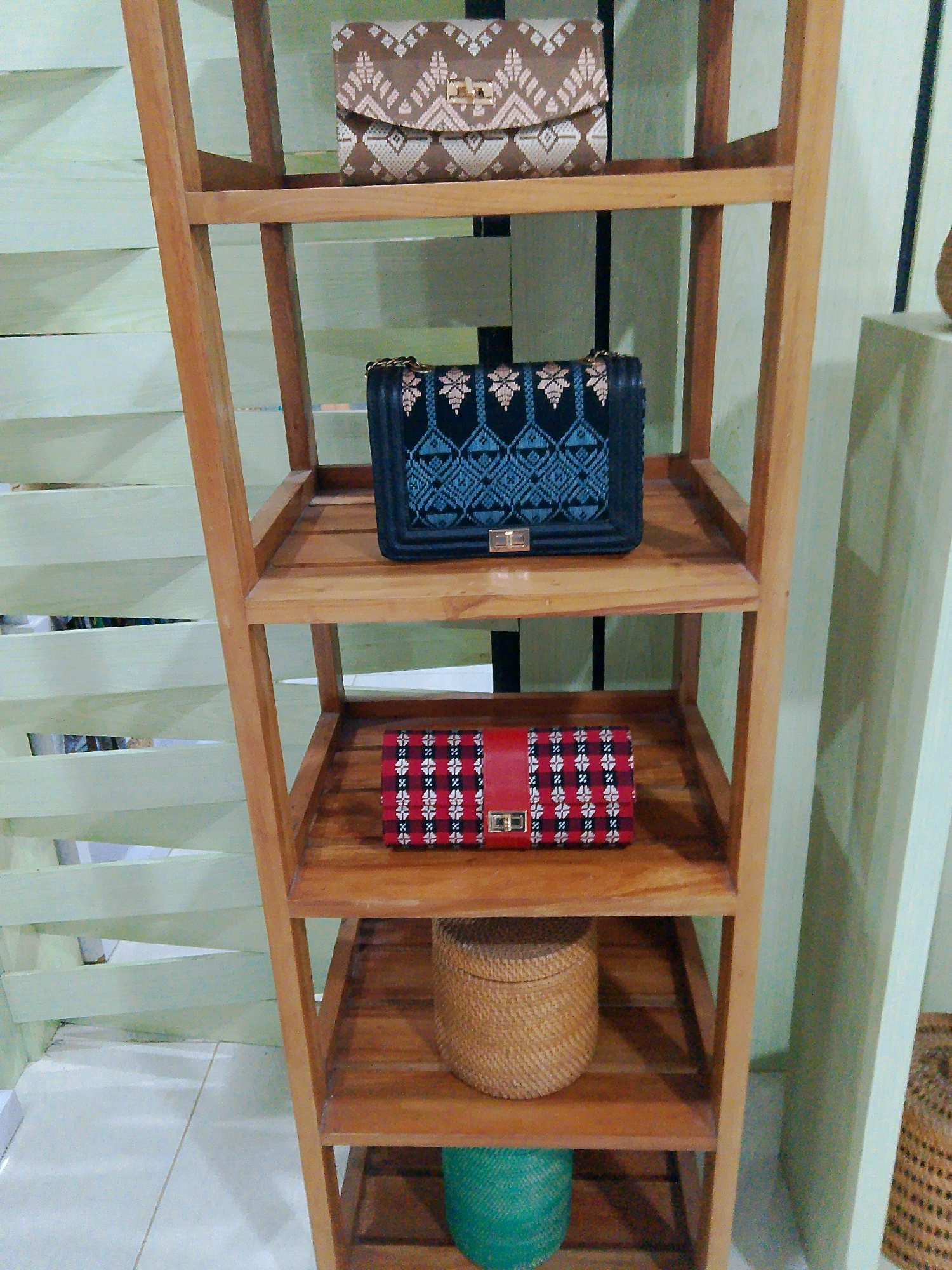 A wooden, multi-tiered shelving unit displays several woven handbags and baskets. The top shelf holds a small, rectangular brown clutch with a geometric woven pattern in light beige. Below it, a black handbag with a black and blue intricate woven pattern and a gold clasp is showcased. The third shelf contains a red and black rectangular clutch with a woven pattern of white floral motifs and a gold clasp. On the fourth shelf, a small, round, light brown woven basket with a lid sits. The bottom shelf is partially visible and appears to hold a green woven spherical object. The shelving unit is placed against a wall with a light green, horizontally slatted background. The floor is tiled in a light color. The scene is set indoors. No people are present. The location is a shop in Bunkate, Indonesia, likely selling handicrafts.