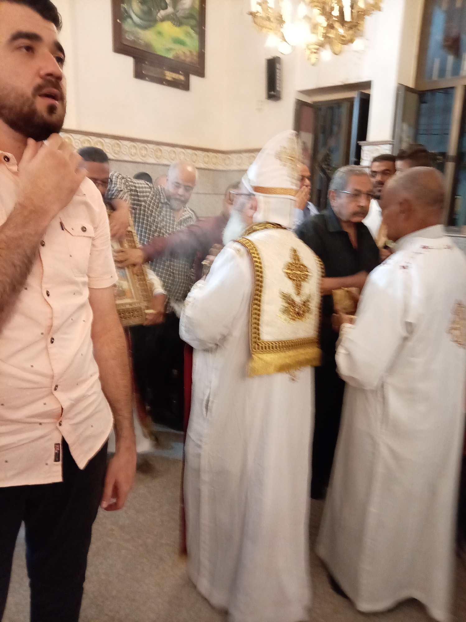 The image shows a group of people inside what appears to be a church. A man in a peach-colored shirt and black pants stands in the foreground, partially obscuring the view. Several men are gathered around a central figure wearing white robes and a tall white headdress adorned with gold. Another man is also wearing similar white robes. Behind them, other individuals are visible, some holding a decorated box. The interior features a painting on the wall, ornate molding, and a chandelier. The location appears to be in Unknown, Unknown.