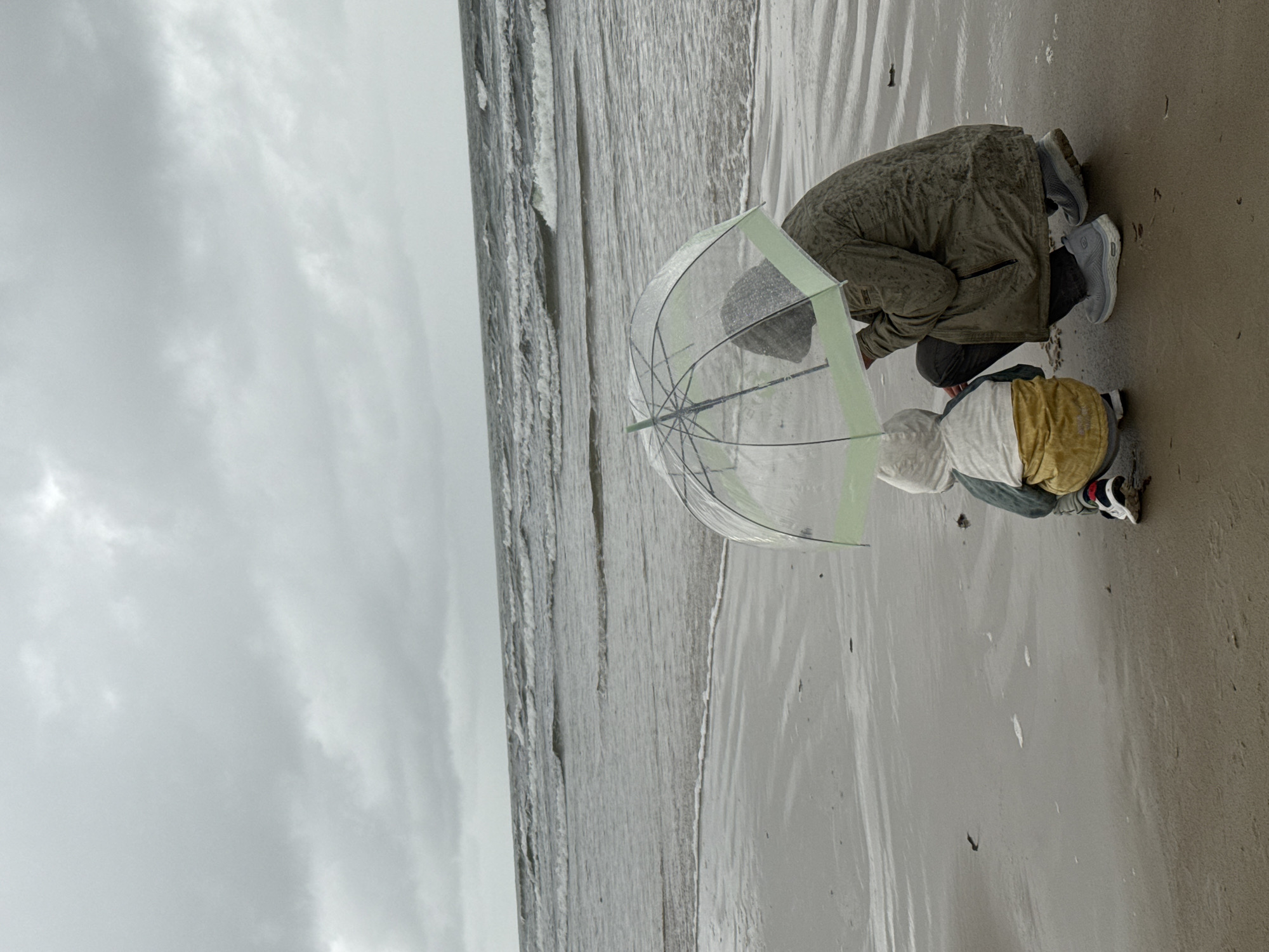 The image captures a somber scene on the beach in Liepaja, Latvia, under an overcast sky. Two individuals, an adult and a child, are crouched on the wet sand, sheltered by a clear umbrella with a light green trim. The adult, wearing a muted green, textured jacket and dark pants, is positioned behind the child. The child, facing away from the camera, is dressed in a hooded jacket with a white and gray top layer and a bright yellow bottom layer, and is wearing sneakers. They are positioned close together, indicating a shared activity or a moment of quiet contemplation. The choppy, gray waves of the Baltic Sea roll onto the sandy shore, suggesting a blustery or rainy day. The overall atmosphere is one of solitude and resilience against the elements.