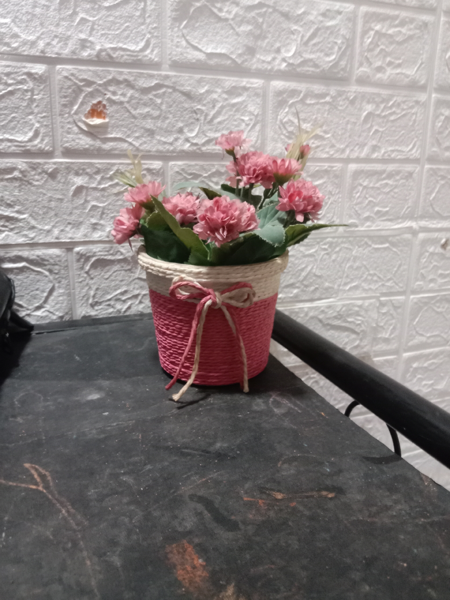 A close-up, slightly tilted shot shows a potted artificial flower arrangement with pink blossoms and green leaves. The pot is a cylindrical container wrapped in twine, with the bottom two-thirds being bright pink and the top portion a natural tan color. A thin tan and pink twine is tied around the pot in a bow. The arrangement is positioned on a dark, textured surface that appears to be a table or shelf. Behind the pot, a white wall with a raised brick pattern serves as the backdrop. A hint of a dark, curved metal object, possibly a railing, is visible on the right side of the frame. There are no people or animals present in the image. The overall lighting is dim, with the focus primarily on the flower pot.