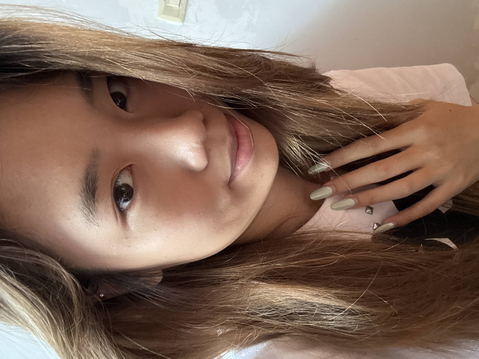 A close-up, forward-facing portrait of a young East Asian woman with long, light brown hair and light skin. She is wearing a pale pink shirt with some black detail visible. Her right hand, with light green nail polish and a silver stud on one fingernail, is resting on her chest, near her neck. She has a small silver stud earring in her left ear. The background is a plain, light-colored wall. The image appears to be taken indoors, possibly in a home or studio environment. There is no identifiable information linking this image to Guandu, China.
