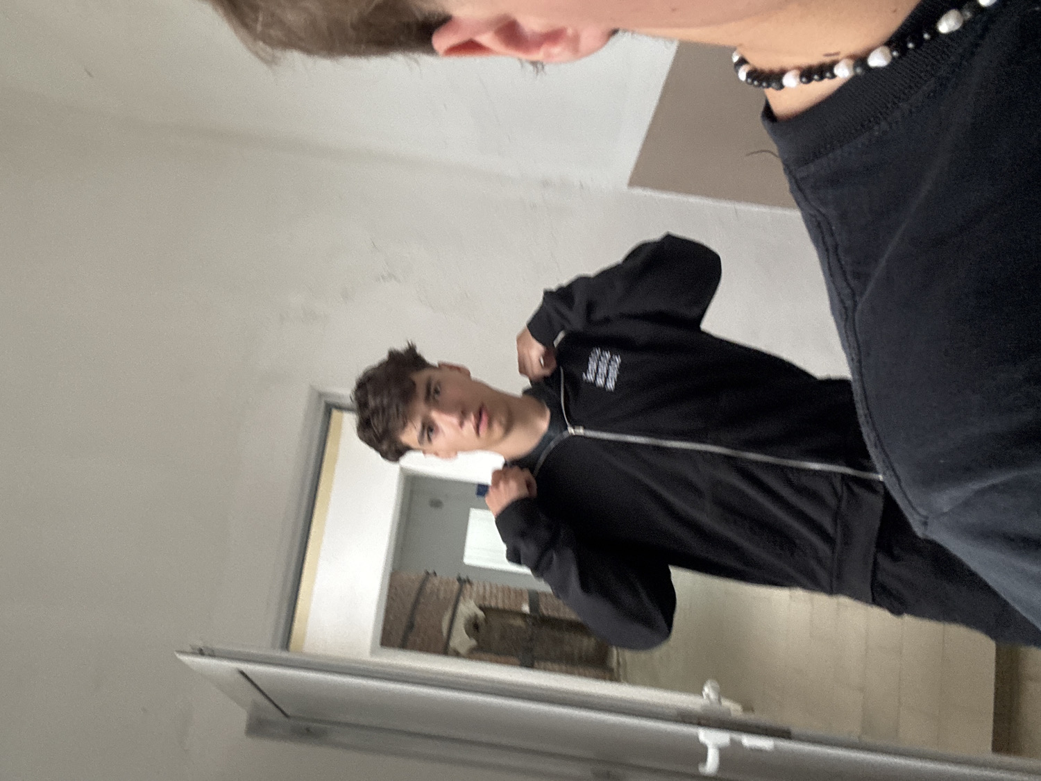 The image captures two young men indoors in what appears to be a hallway or room, possibly in Troyan, Bulgaria. The perspective suggests a selfie or a photo taken in a mirror, as the top of the frame shows the head (forehead and ear) and shoulder of one person, who is wearing a black top and a beaded necklace composed of alternating black and white spherical beads.
The main subject, a young man with dark, styled hair, is positioned in the mid-ground, looking directly towards the camera with a neutral expression. He is wearing a black zippered hoodie or jacket, which features a prominent silver zipper down the front. On the left chest area of his hoodie, there is a multi-lined white text or logo that is partially obscured and difficult to identify clearly. His hands are near the collar or hood of his jacket.
The immediate background consists of plain white walls. Through an open doorway or reflection on the left, a glimpse of another room or area is visible, containing some reddish-brown elements that resemble brickwork or wooden structures, hinting at an older interior design. The lighting suggests it is daytime. The image itself is rotated at an angle.