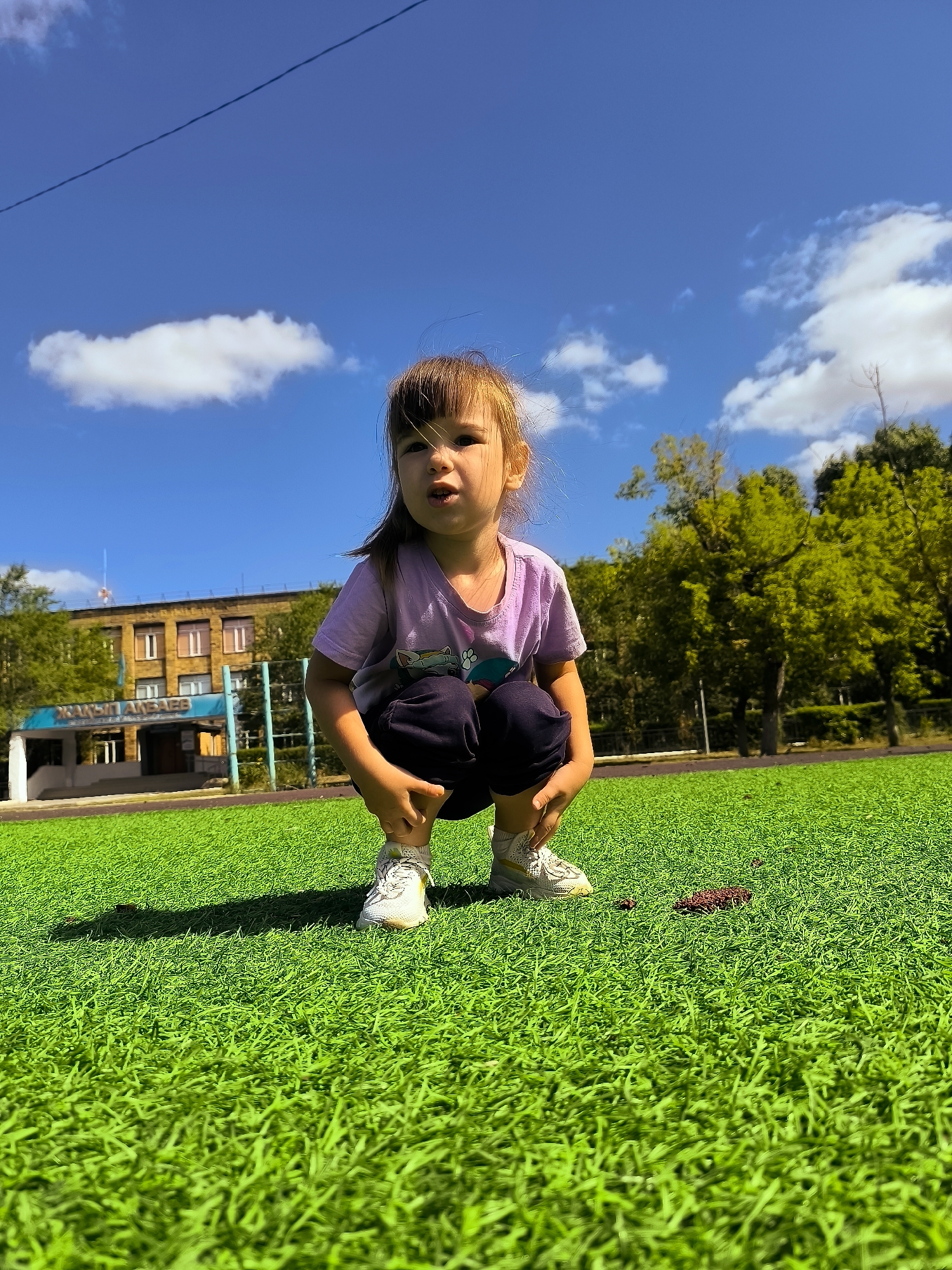 A young girl wearing a purple t-shirt and dark pants squats on a vibrant green artificial turf field under a bright blue sky with scattered white clouds. She is positioned in the foreground, facing slightly to her right, with her hands resting on her knees. Behind her, a light brown building with multiple windows is visible, and to the right, lush green trees form a backdrop. A blue sign with white lettering is mounted on the building's facade. The scene appears to be an outdoor sports complex or school grounds in Qarağandı city administration, Kazakhstan, indicated by the building and the athletic field.