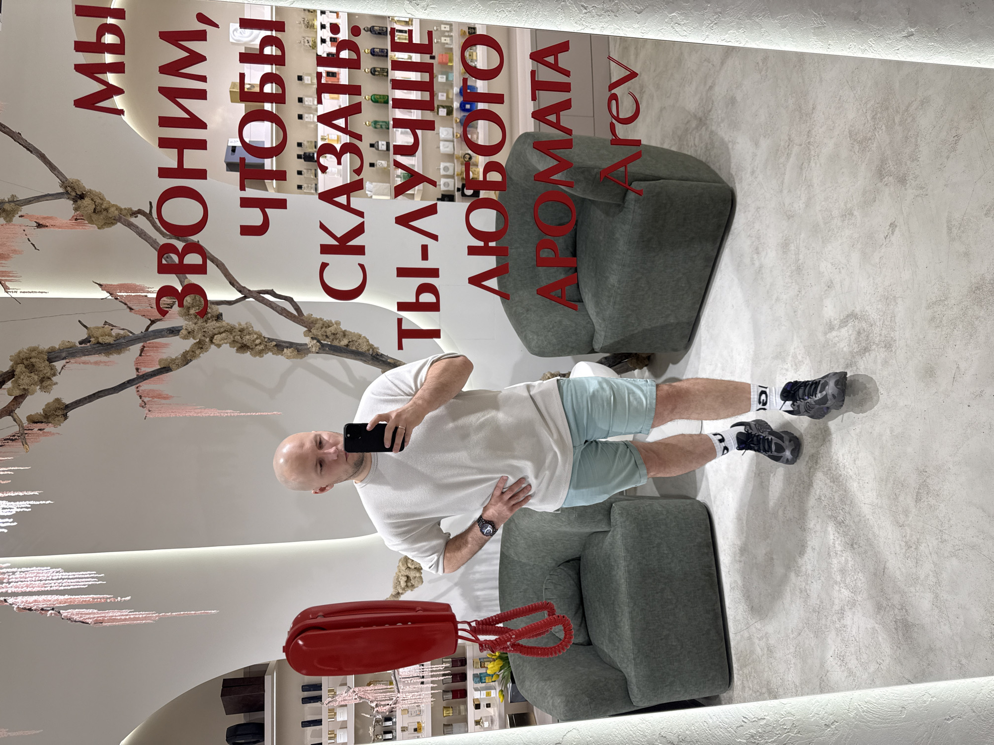 The image is a mirror selfie taken by a bald man standing in a retail store. He is wearing a light-colored textured t-shirt, light blue shorts, white socks with black markings, and dark athletic shoes. He is holding a smartphone in his right hand and has his left hand on his hip.
The background of the mirror reflection shows shelves filled with bottles, likely perfumes, and two upholstered armchairs. A red rotary-style telephone is mounted on the wall to the left. Red text is displayed on the mirror, with the visible portion reading "МЫ ЗВОНИМ, ЧТОБЫ СКАЗАТЬ: ТЫ-ЛУЧШЕ ЛЮБОГО АРОМАТА Arev". This translates to "WE CALL TO SAY: YOU ARE BETTER THAN ANY FRAGRANCE Arev".
The store appears to be well-lit and modern, with a branch-like decorative element hanging from the ceiling. The floor is a light-colored concrete. The overall context suggests the image was taken in a perfume boutique or a similar retail establishment. There is no information in the image itself that indicates a specific location in the city of Krasnoyarsk, Russia.