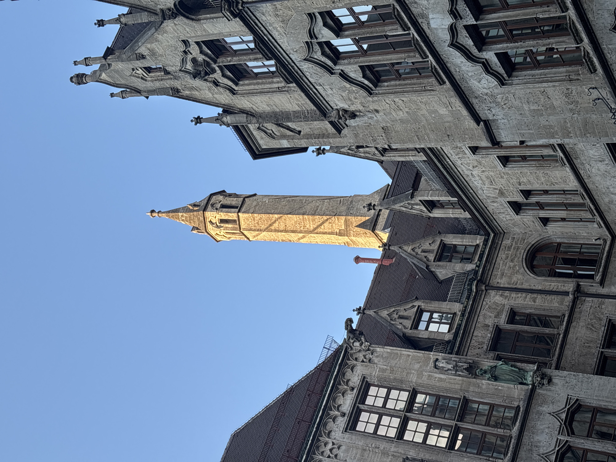 The image shows a low-angle view of the Neues Rathaus (New Town Hall) in Munich, Germany, on a clear day. The architecture is Gothic Revival, characterized by intricate stone carvings, pointed arches, and steep roofs. A tall, slender tower with a spired roof is visible in the background, bathed in sunlight, contrasting with the cooler tones of the adjacent buildings. Gargoyles and statues adorn the facade of the Neues Rathaus, adding to its historical and ornate appearance. The sky is a vibrant, cloudless blue. The scene is static and shows no people or animals.