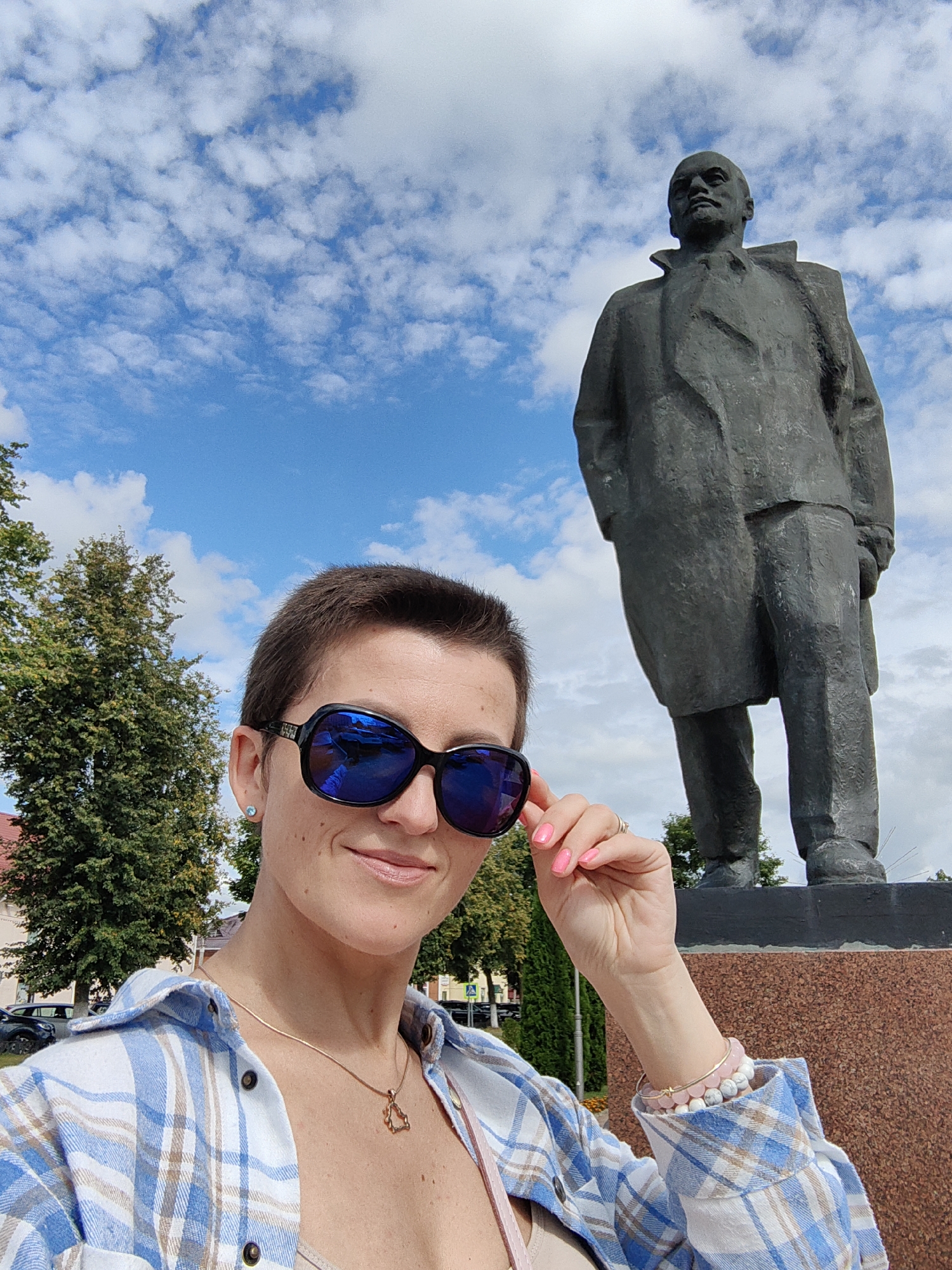 The image is a selfie taken by a woman with short brown hair, wearing large blue-tinted sunglasses and a blue and white plaid shirt. She has bright pink fingernails and is wearing a delicate gold necklace. She is standing outdoors in front of a large, dark-colored statue of a man in a coat, positioned on a brown stone pedestal. Behind the statue, there are green trees and a blue sky with white clouds. The woman is touching her sunglasses with her right hand. The statue is identifiable as Vladimir Lenin and is located in the city of Pastavy, Belarus. The background shows buildings and parked cars, indicating an urban setting.