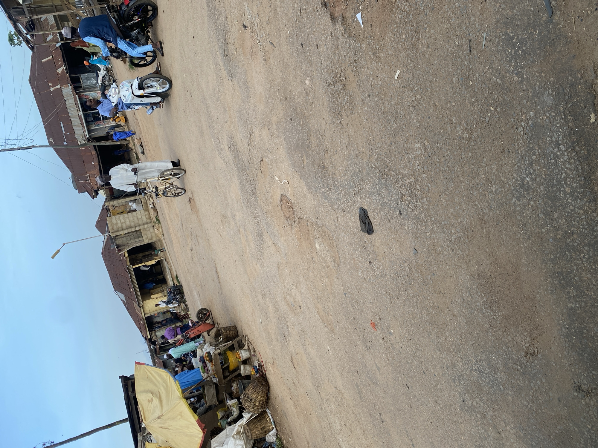 The image depicts a street scene in Jos, Nigeria, characterized by an unpaved road with visible potholes and scattered debris, including a black sandal and what appears to be an orange wrapper. On the left side of the frame, a market stall is set up under a yellow awning, displaying various goods, including fruits and baskets, with several individuals present. Further down the street, three men are visible. One man, dressed in a white traditional garment and cap, stands next to a small bicycle. Another man, in a blue shirt, rides a white motorcycle, while a third man, wearing a light blue outfit and cap, sits on a black motorcycle. Behind them, there are several structures with corrugated iron roofs, indicative of a typical marketplace or residential area in Jos. The sky is overcast, suggesting a cloudy day.