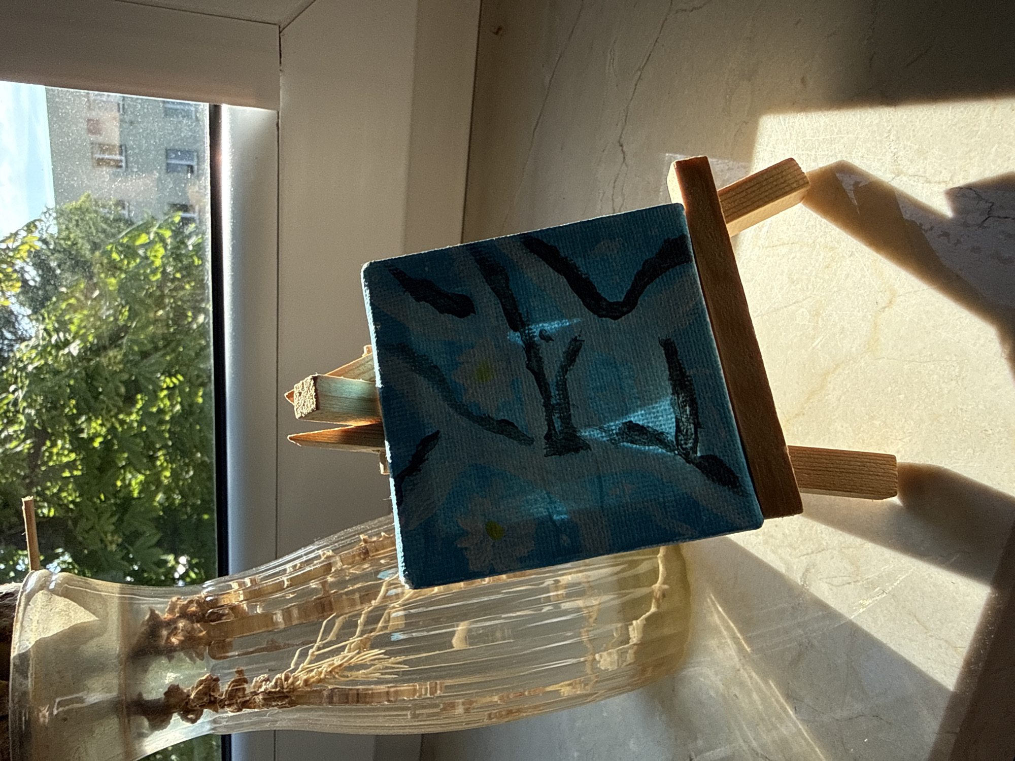 The image shows a small, square painting on a wooden easel placed on a windowsill. The painting is abstract, featuring a blue background with dark, swirling lines and some small white shapes that might represent flowers. To the left of the painting is a clear glass vase containing dried plant material with visible roots and stems. Sunlight streams in from a window, casting shadows on the windowsill and the wall behind it. Through the window, a building with multiple windows and some green foliage can be seen. The scene is likely an indoor setting, possibly a home or studio, with a view of the urban environment of Łódź, Poland. No people or specific actions are depicted.