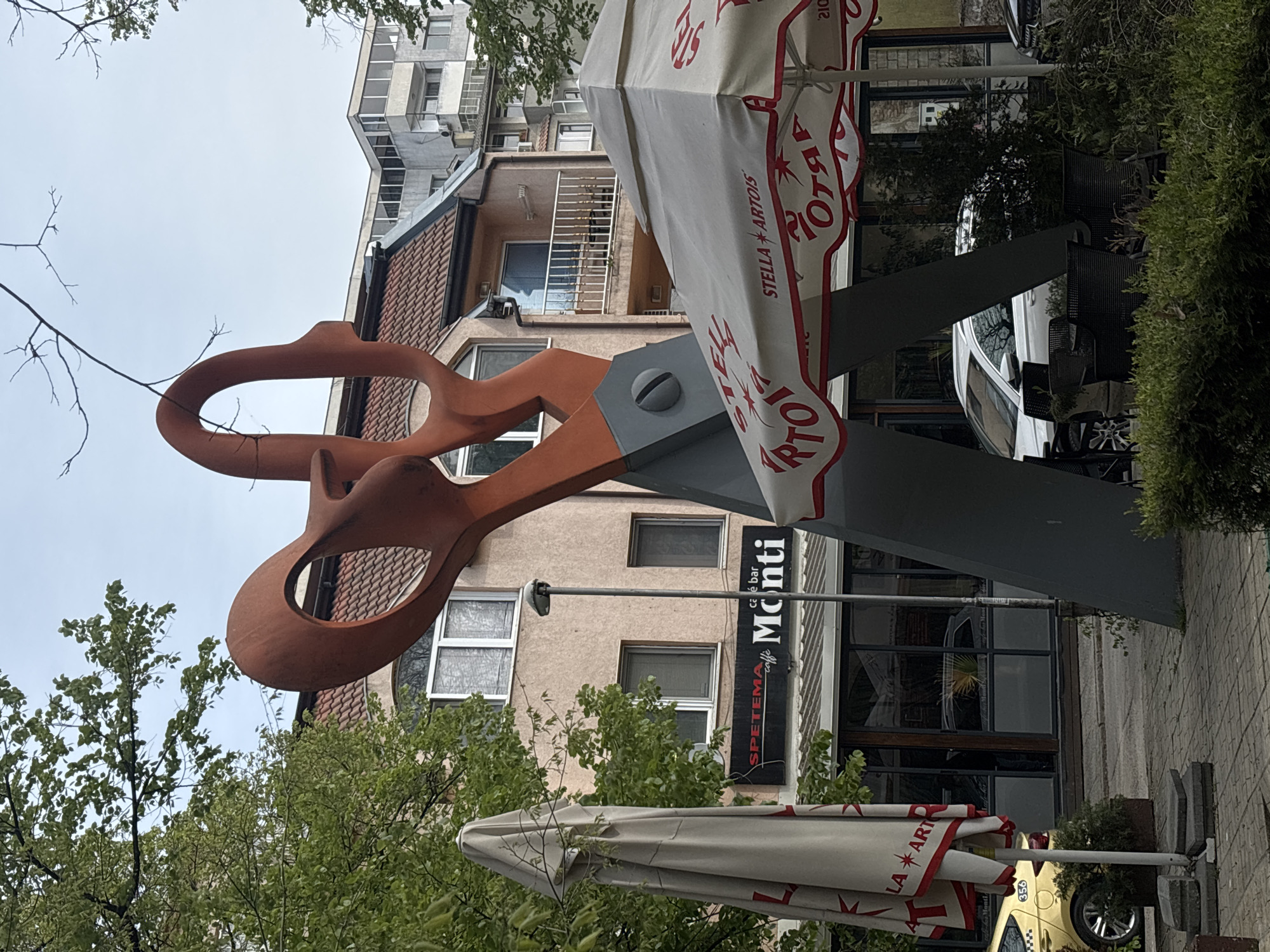 Here is a description of the image:
The image captures an outdoor scene in Varna, Bulgaria, prominently featuring a large, possibly metallic, sculpture of a pair of open scissors. The scissors are painted in a rust-like color and are mounted on a gray pole or stand. In the background is a multi-story building with light-colored facades and various windows.
A sign for "SPETEMA caffe bar Monti" is visible, suggesting the location is near a cafe or bar called "Monti." There are also partly visible outdoor umbrellas branded with "STELLA ARTOIS" lettering, indicating a likely seating area. Elements like trees, a glimpse of a yellow taxi, and other indistinct building details fill the background, creating an urban atmosphere. The sky is overcast.