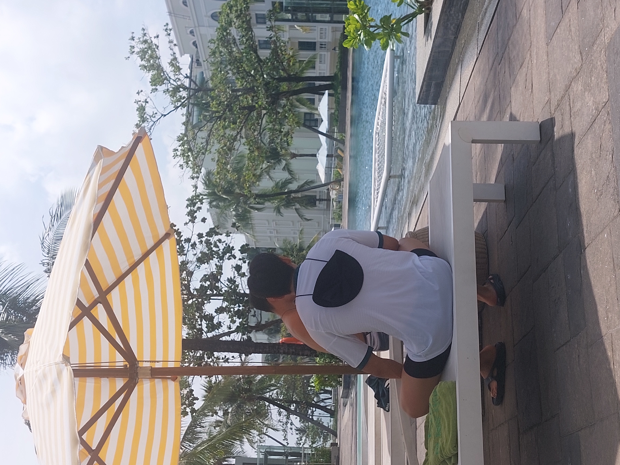 Two individuals are seated on a white lounge chair under a yellow and white striped umbrella. The person in the foreground is wearing a white t-shirt and black shorts, with a black face mask draped over the back of their neck. They are facing away from the camera, towards a swimming pool. The second individual, partially visible behind the first, appears to be wearing swim trunks. The scene takes place outdoors, likely at a resort in Phú Quốc, Vietnam, given the presence of palm trees and the architectural style of the buildings in the background, which resemble a modern hotel or resort complex. The swimming pool features a floating water hammock or mat. The ground is paved with stone tiles. The overall impression is a relaxed, sunny day by a pool.