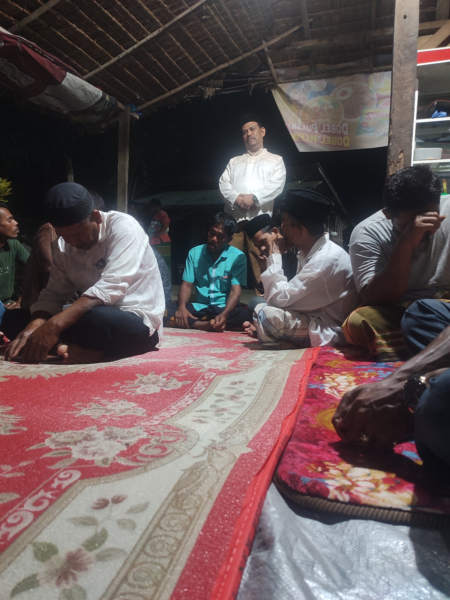 The image depicts a gathering of men in what appears to be an open-air structure with a thatched roof. The setting is dimly lit, suggesting it is either evening or indoors with minimal lighting. Several men are seated on a large, patterned rug on the ground. One man in the foreground, wearing a white shirt and a black cap, is looking down at his hands. To his right, another man in a grey t-shirt is also seated, with his head bowed. In the background, a man in a white traditional outfit and a black cap stands with his hands clasped. Another man in a teal shirt is seated to the left. A banner with text in what appears to be a foreign language is visible hanging behind the standing man. The environment suggests a casual or informal gathering. The location is identified as Langkuta, Indonesia.