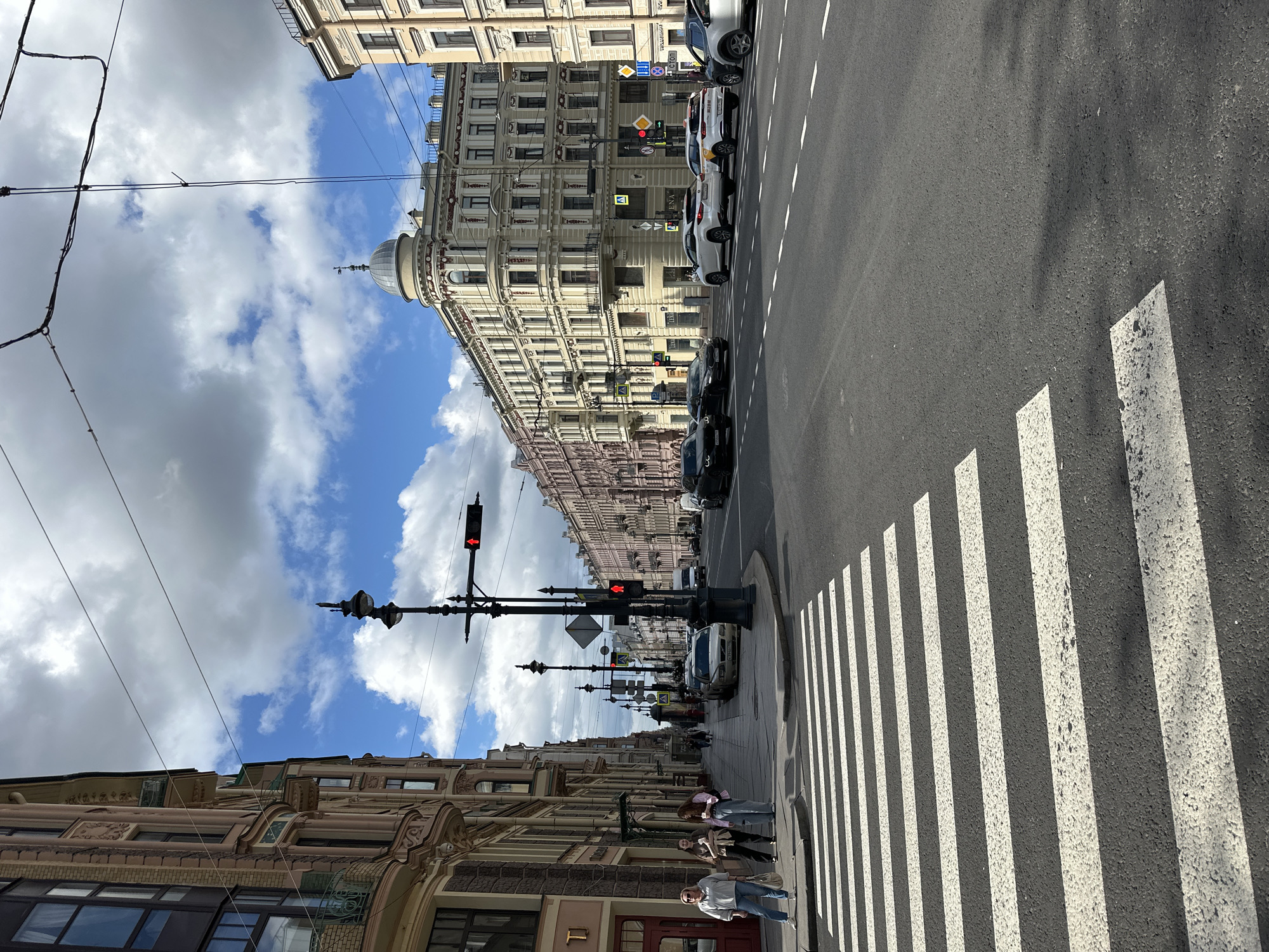 A street scene in Saint Petersburg, Russia, shows a wide asphalt road with a pedestrian crossing in the foreground. On the left, a tall, ornate building with intricate architectural details and balconies lines the sidewalk. Several people are present on the sidewalk, including two women walking and another two standing near the crossing. A black lamppost with multiple fixtures and traffic lights stands prominently near the crossing. Across the street, a series of multi-story buildings with detailed facades stretches into the distance, some appearing to be apartment buildings or commercial spaces. Cars are visible on the road, some driving and others parked along the side. The sky is partly cloudy, with patches of blue sky visible between white cumulus clouds. The overall impression is a typical urban streetscape on a day with mixed weather conditions.