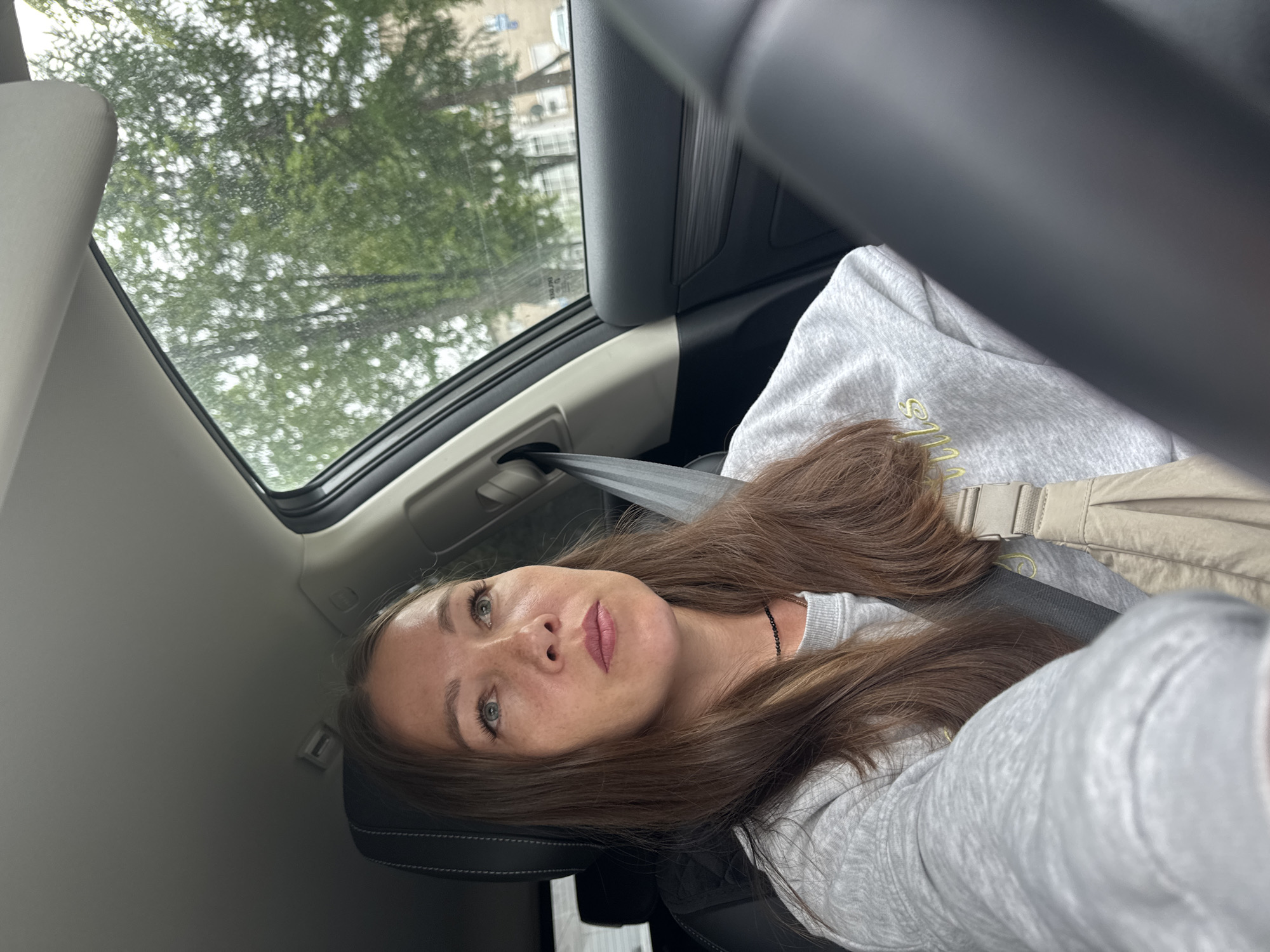 A medium shot shows a woman in the driver's seat of a car, looking upward and to the right. The car's interior is visible, including the steering wheel, dashboard, and seatbelt. Outside the car, through the window, trees and buildings can be seen, suggesting an urban environment. The weather appears overcast. The woman has long, brown hair and is wearing a light gray sweatshirt and a dark necklace. A light-colored bag is slung across her shoulder. The scene is captured in daylight.