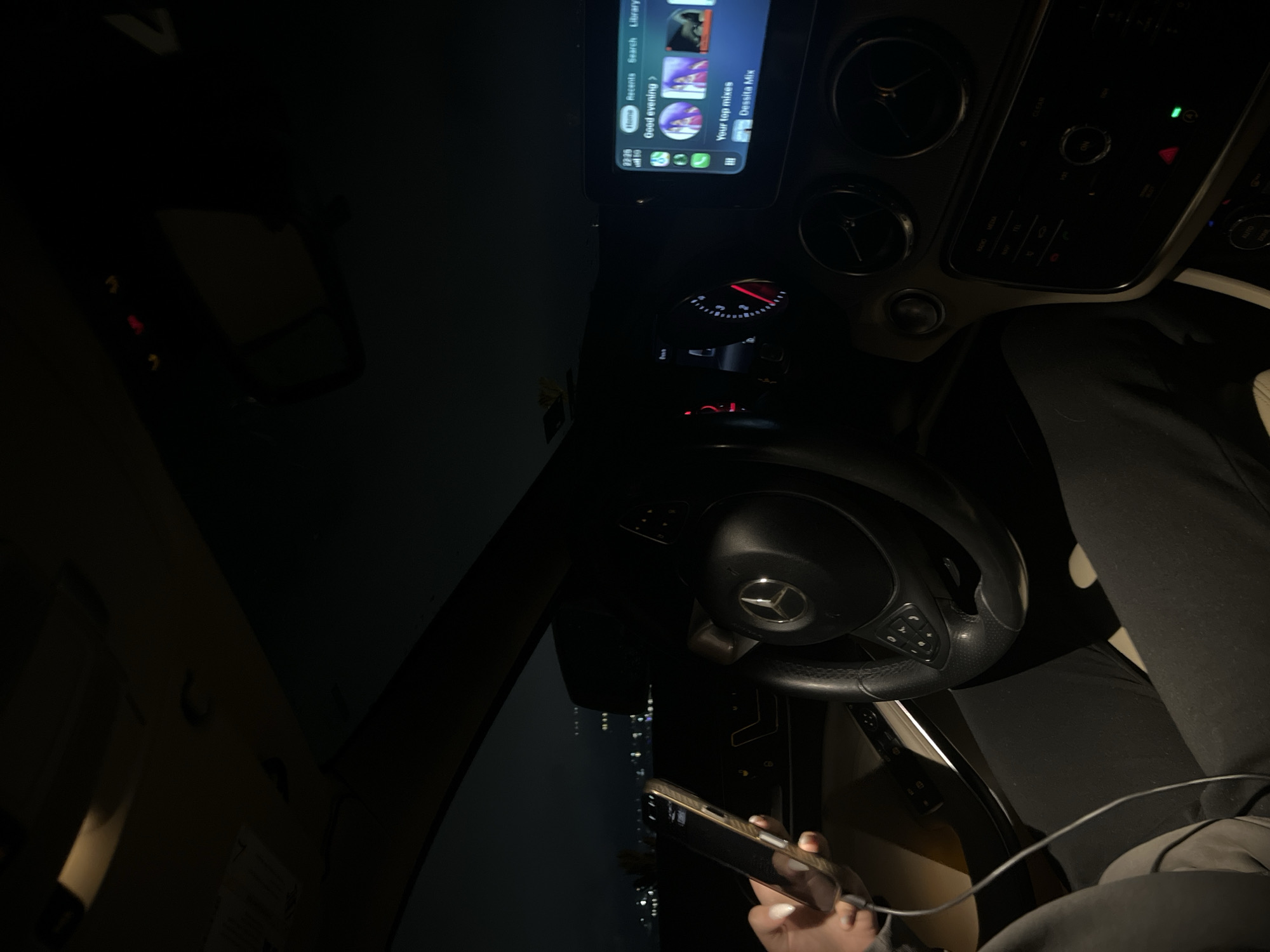The image shows the interior of a car, specifically the driver's area, at night. The steering wheel features a Mercedes-Benz logo, suggesting the car make. The driver is holding a smartphone, which is connected to the car with a cable. The infotainment screen displays "Good evening," and a list of music. The city lights can be seen dimly through the windshield. The location is in a city, identifiable by the skyline, but it can not be ascertained that the exact location is Blagoevgrad, Bulgaria based on the given image.