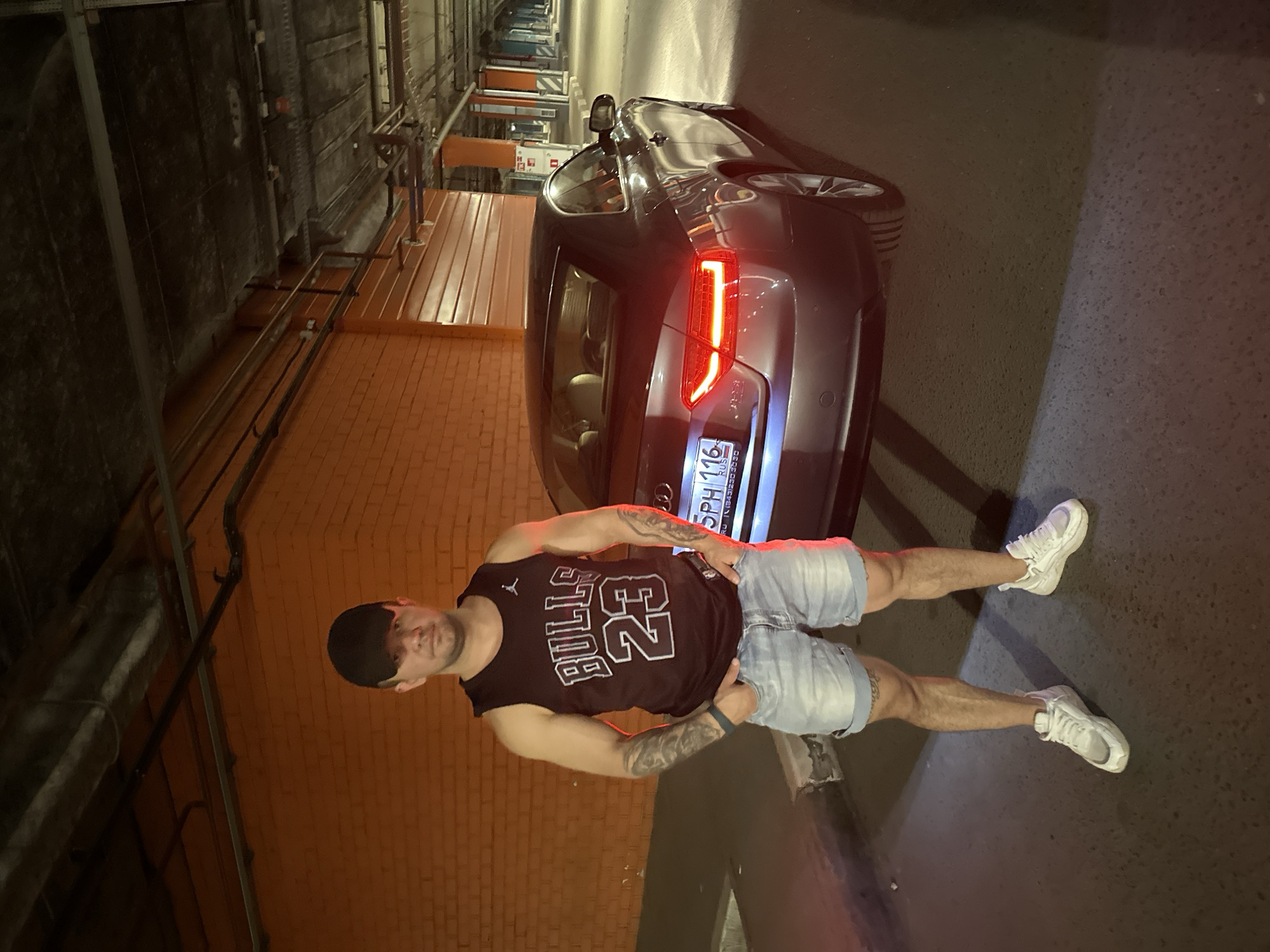 A man wearing a black baseball cap, a dark sleeveless shirt with "BULLS 23" printed on it, and light blue denim shorts stands with his hands in his pockets in front of a dark gray Audi coupe in what appears to be a parking garage. The car's taillights are illuminated. The walls of the garage are orange brick and corrugated metal. The floor is asphalt. The scene is lit by artificial light, casting shadows and creating a warm glow. The license plate of the car is visible and reads "5PH 116". The image captures a casual moment in an urban environment.